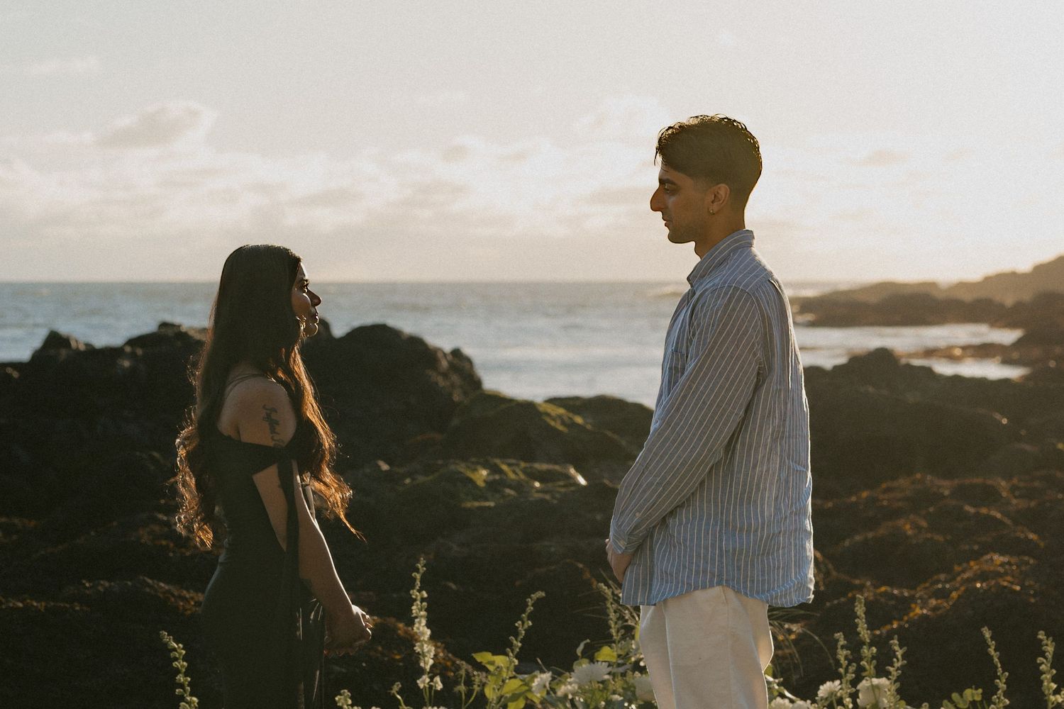 A couple holds hands facing each other on rocky coastal cliffs at golden hour with ocean waves in the background.