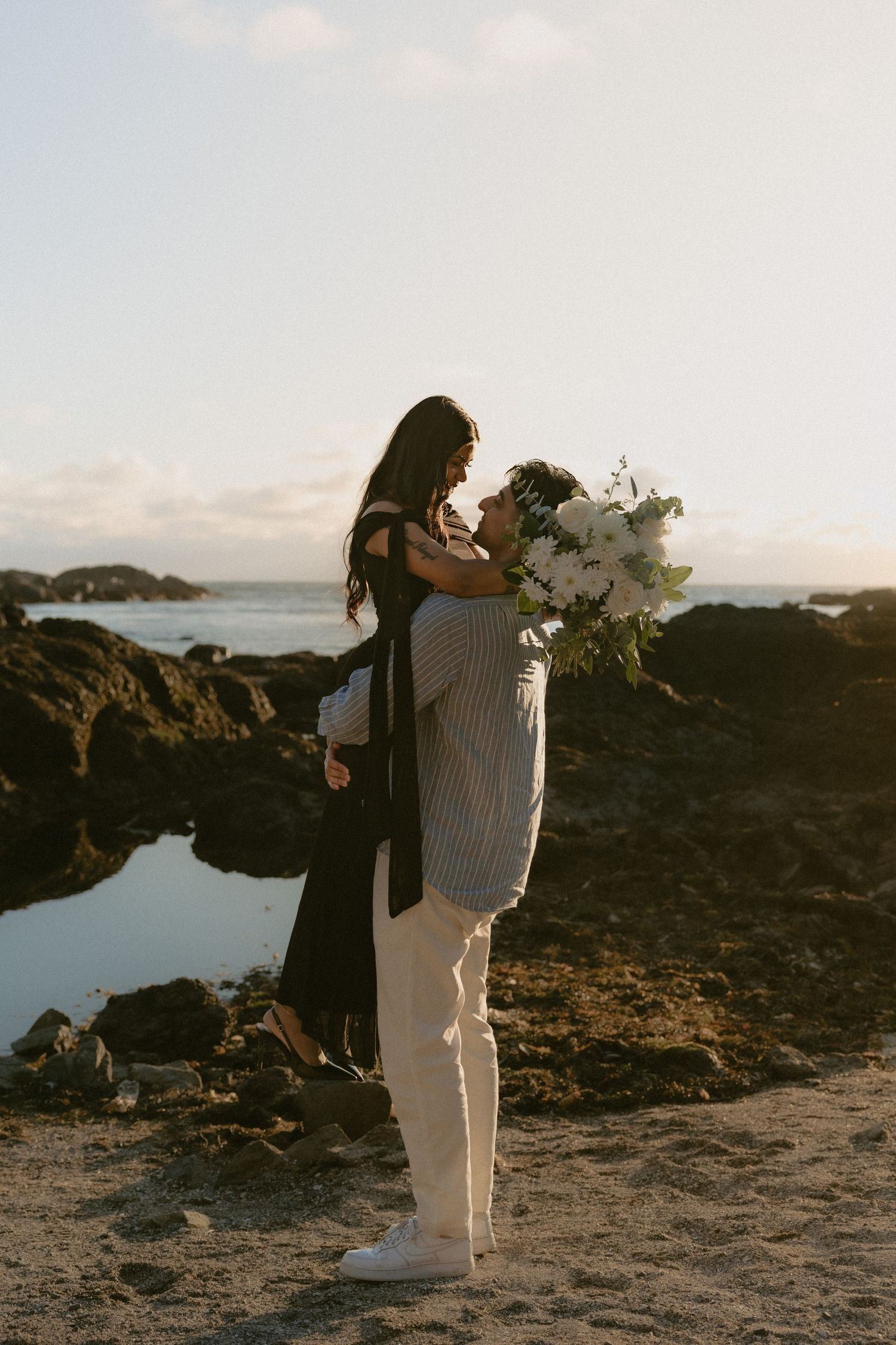 A couple shares a romantic moment on a rocky beach, man lifting woman holding white bouquet at golden hour.
