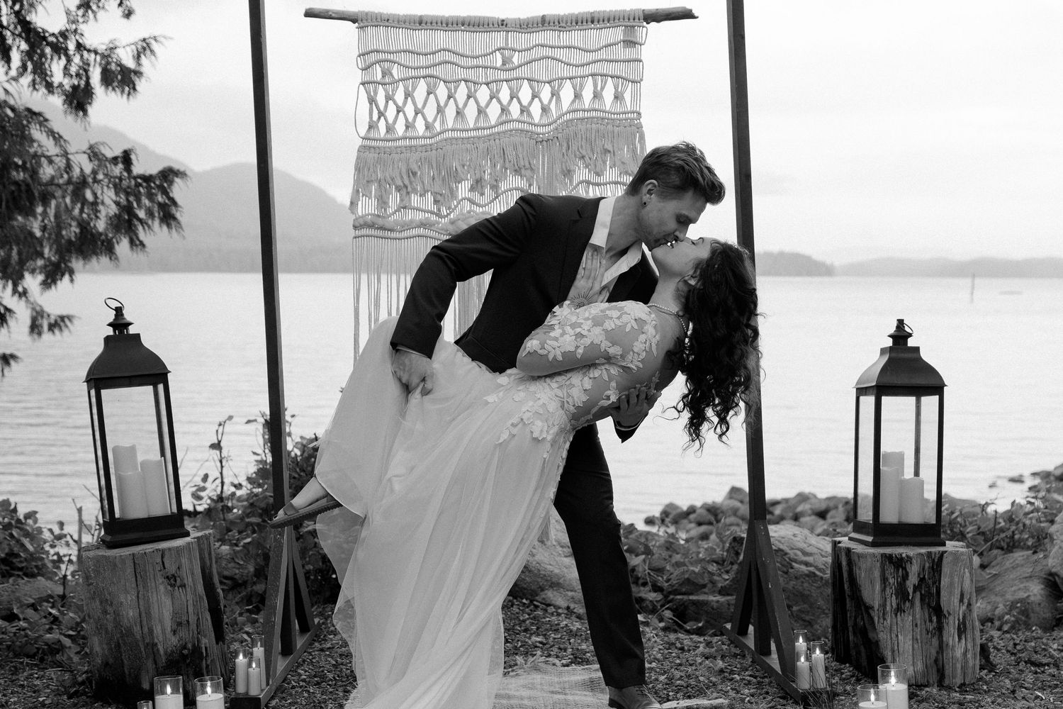 Black and white photo of bride and groom sharing a romantic dip kiss at outdoor waterfront wedding ceremony with lanterns.