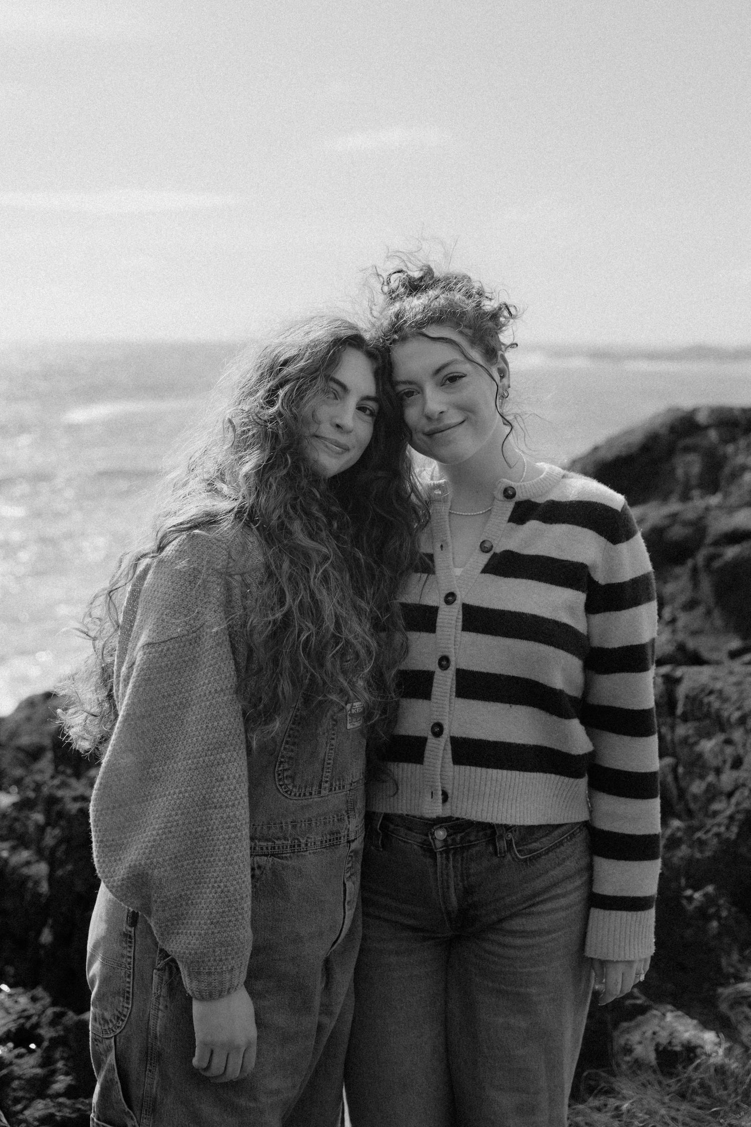 Two young women with curly hair pose together on rocky ocean shore in black and white photo.