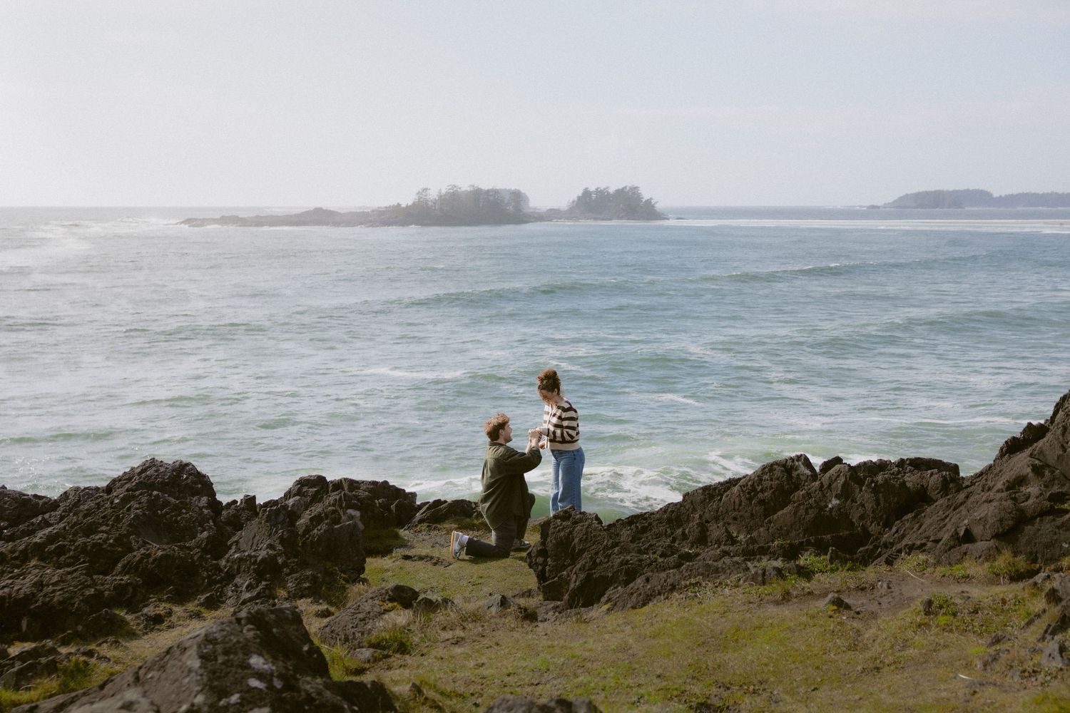 Couple sharing a marriage proposal moment on rocky shoreline with misty ocean and islands behind.