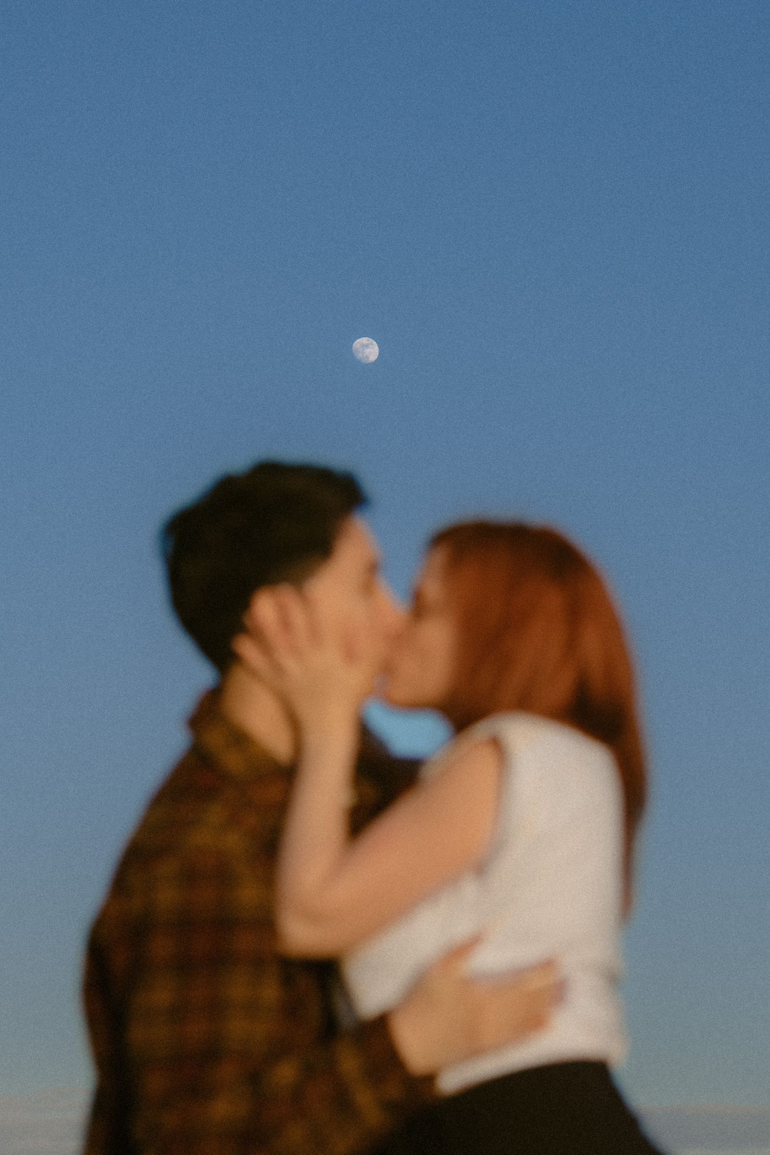 Couple sharing a romantic kiss outdoors under a clear blue sky with a visible moon.