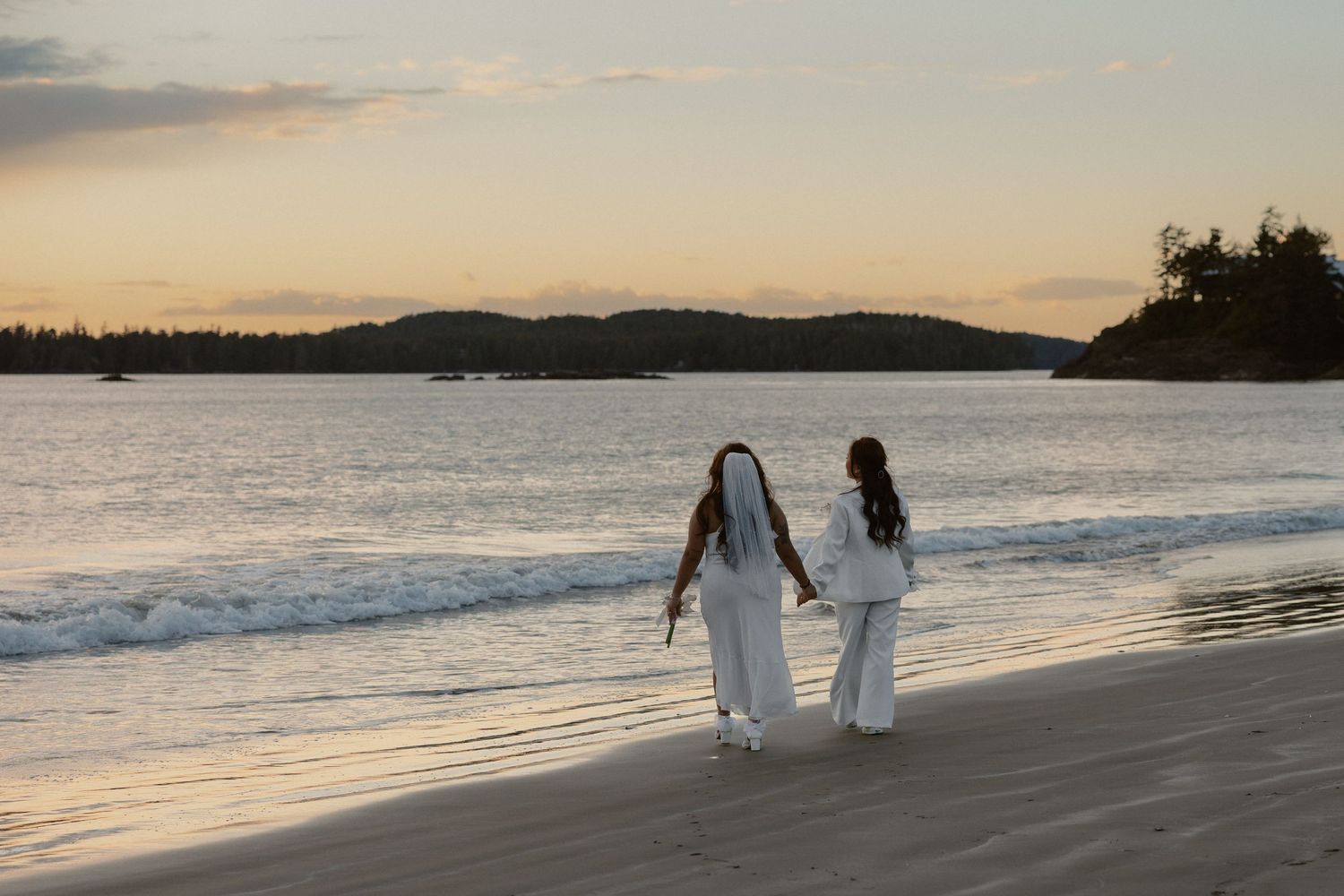 Two people in flowing white attire stroll together on a beach during golden hour.