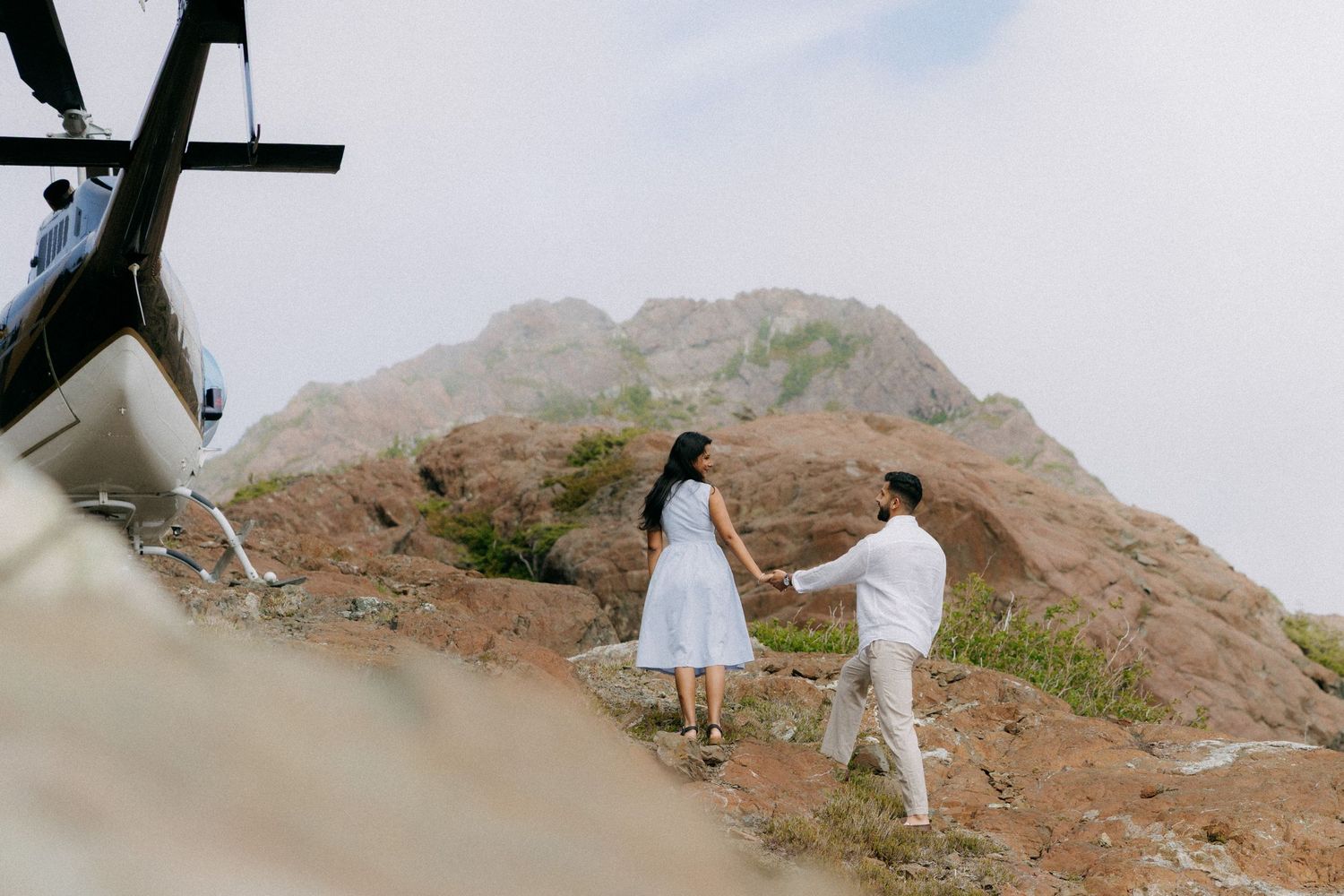 Couple holding hands near a helicopter on a rocky mountain summit during an adventurous engagement photo session.