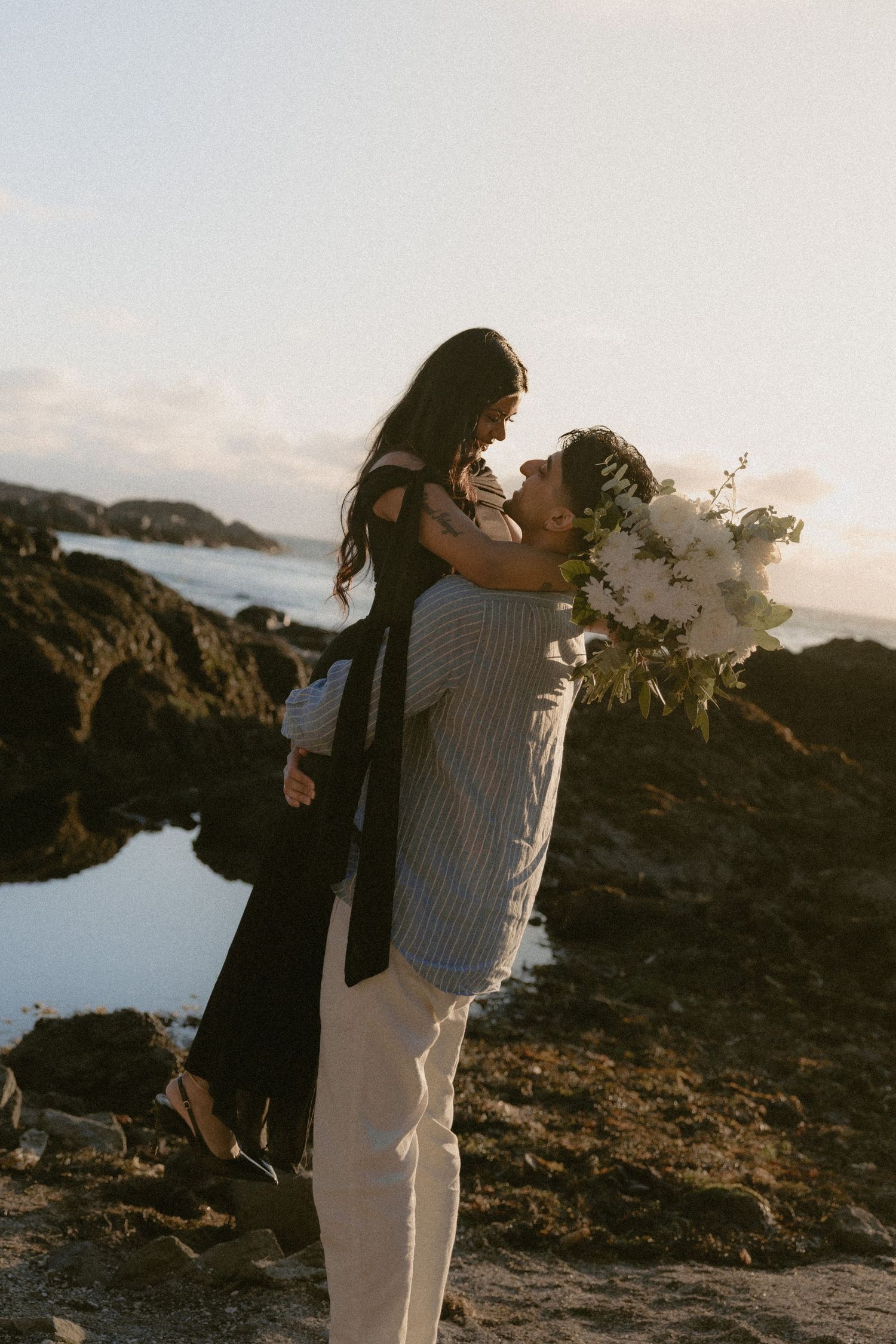 Couple sharing a romantic moment at rocky coastline, man lifting woman holding white floral bouquet at sunset.