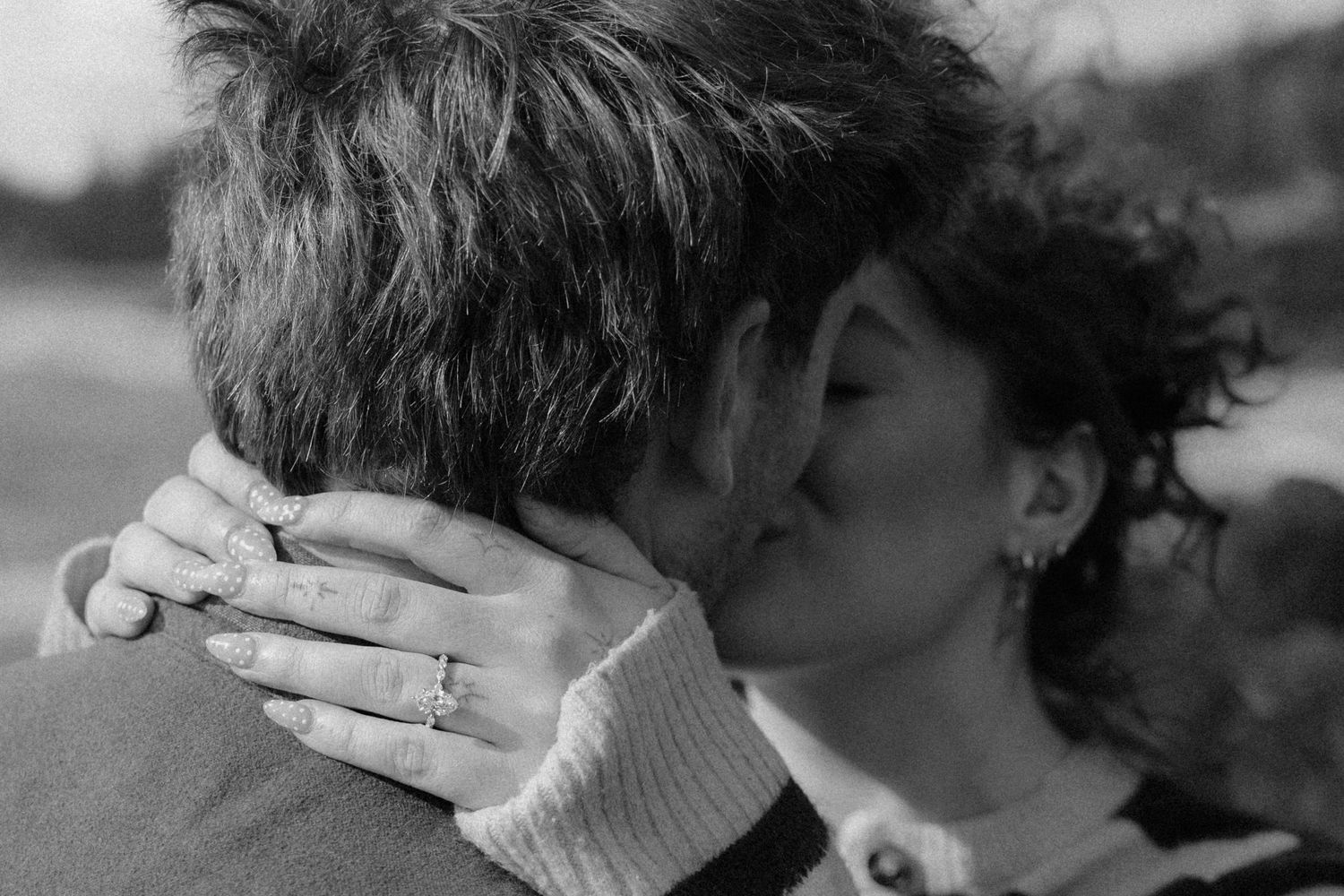 Black and white close-up of a couple sharing a kiss, showcasing an engagement ring on her hand.