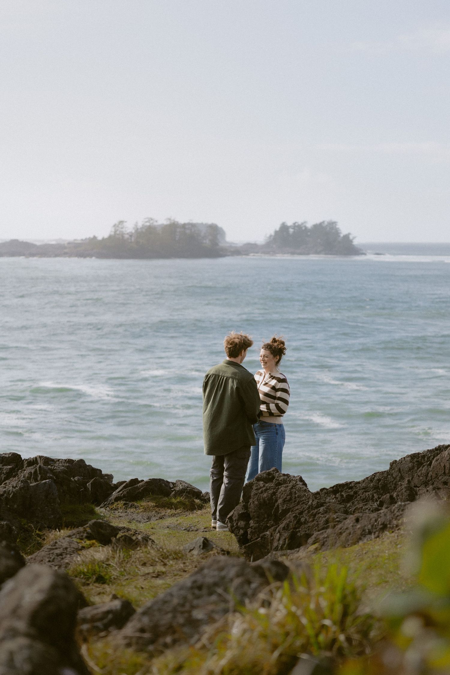 Couple standing on rocky coastal cliffs embracing while overlooking a calm ocean with small islands in the distance.