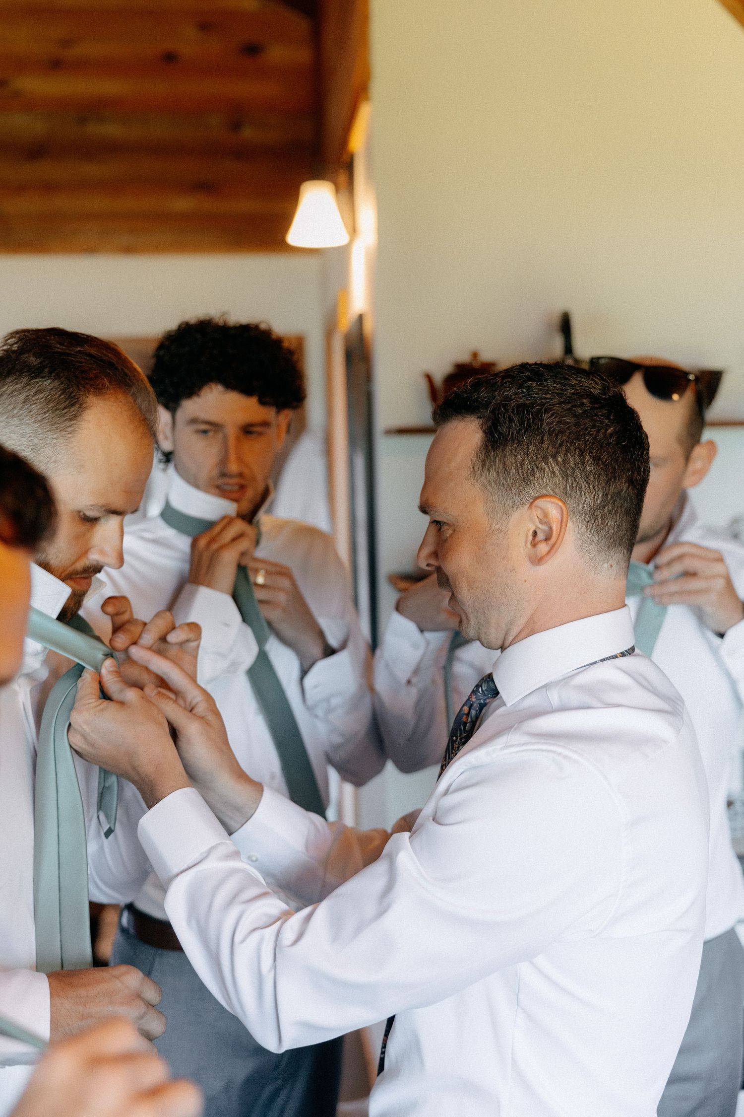 Groomsmen in white shirts help each other tie sage green ties while getting ready for a wedding ceremony.