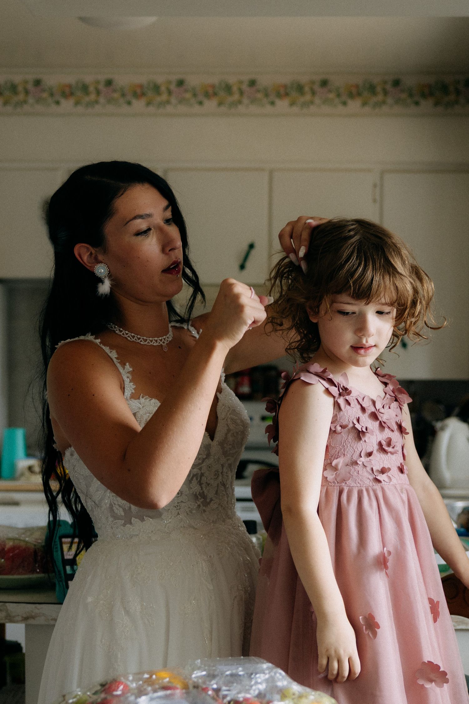 Bride in white lace gown styling flower girl's hair in pink floral dress during wedding preparations.