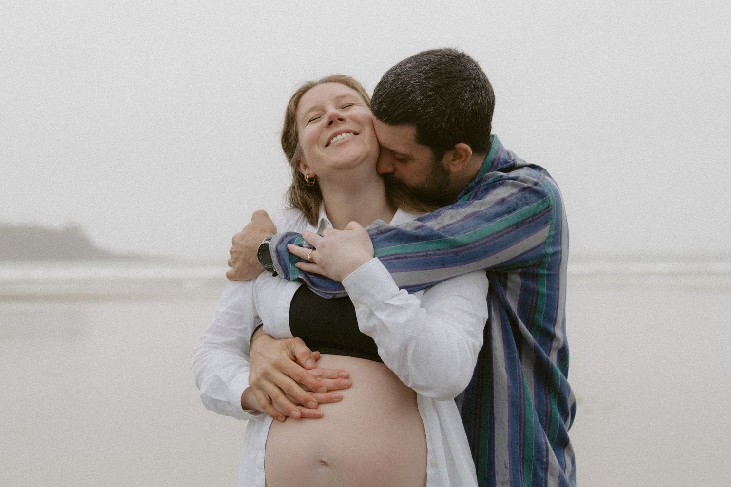 Intimate moment between couple embracing on misty beach during maternity photoshoot.