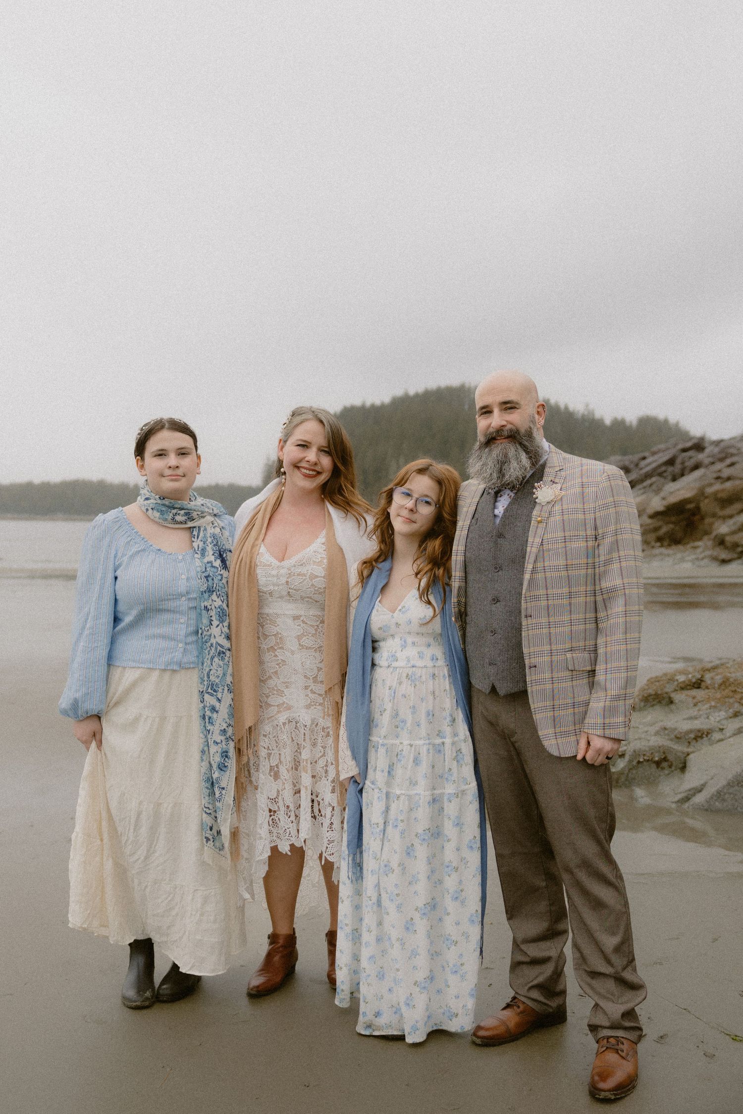 A group poses together on a misty beach wearing vintage-style dresses and clothing in soft blue and beige tones.