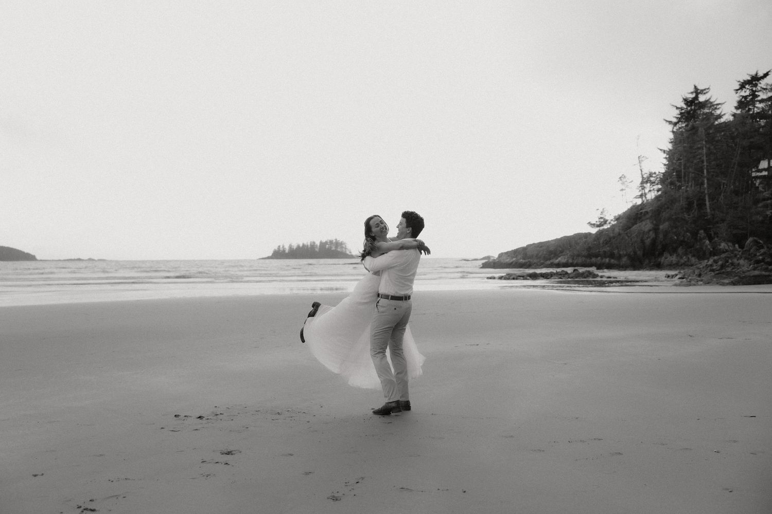 Bride and groom twirling on a misty beach with pine trees in the distance in Tofino