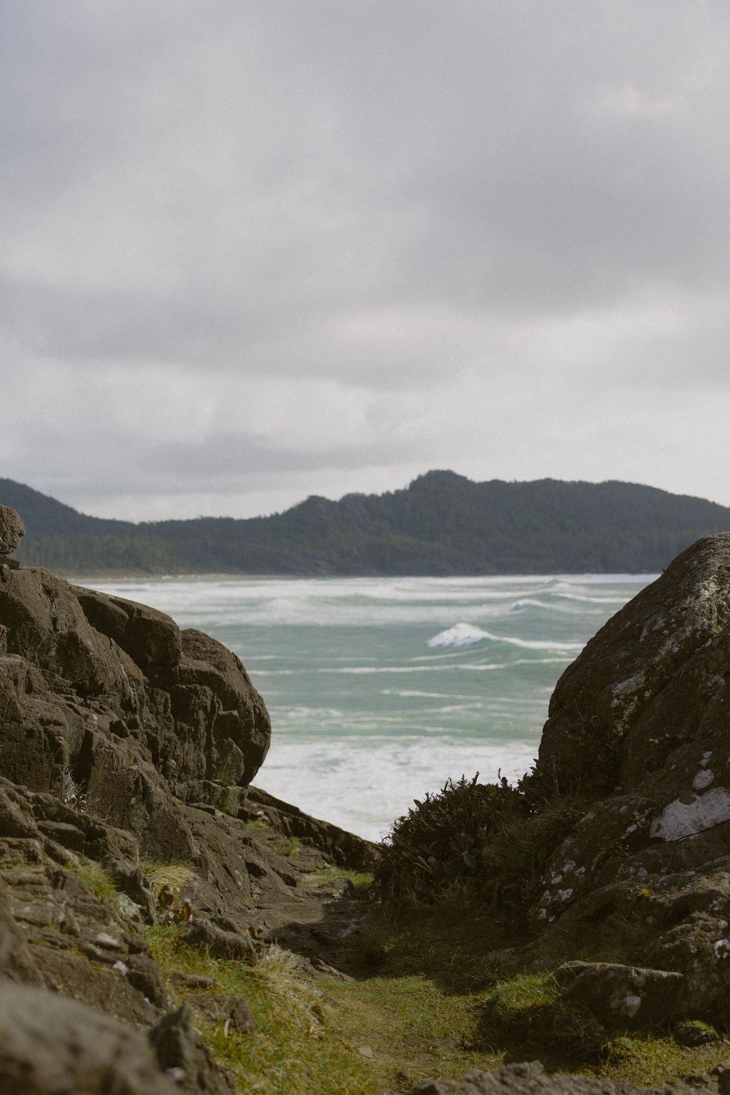 Rocky coastal gap frames churning turquoise waves with forested hills under overcast skies.