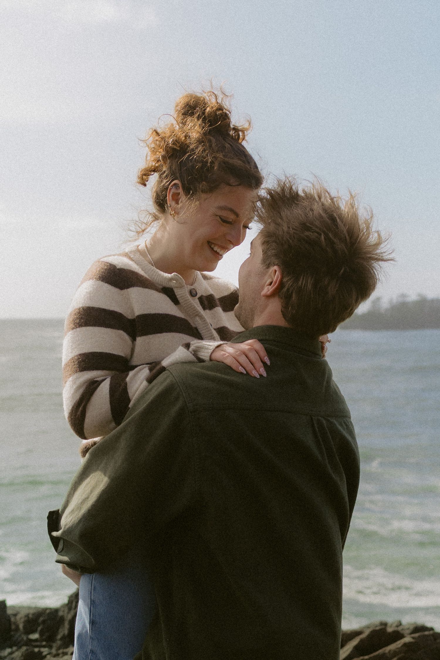 Romantic couple embracing and smiling at each other on rocky seaside cliffs on sunny day.