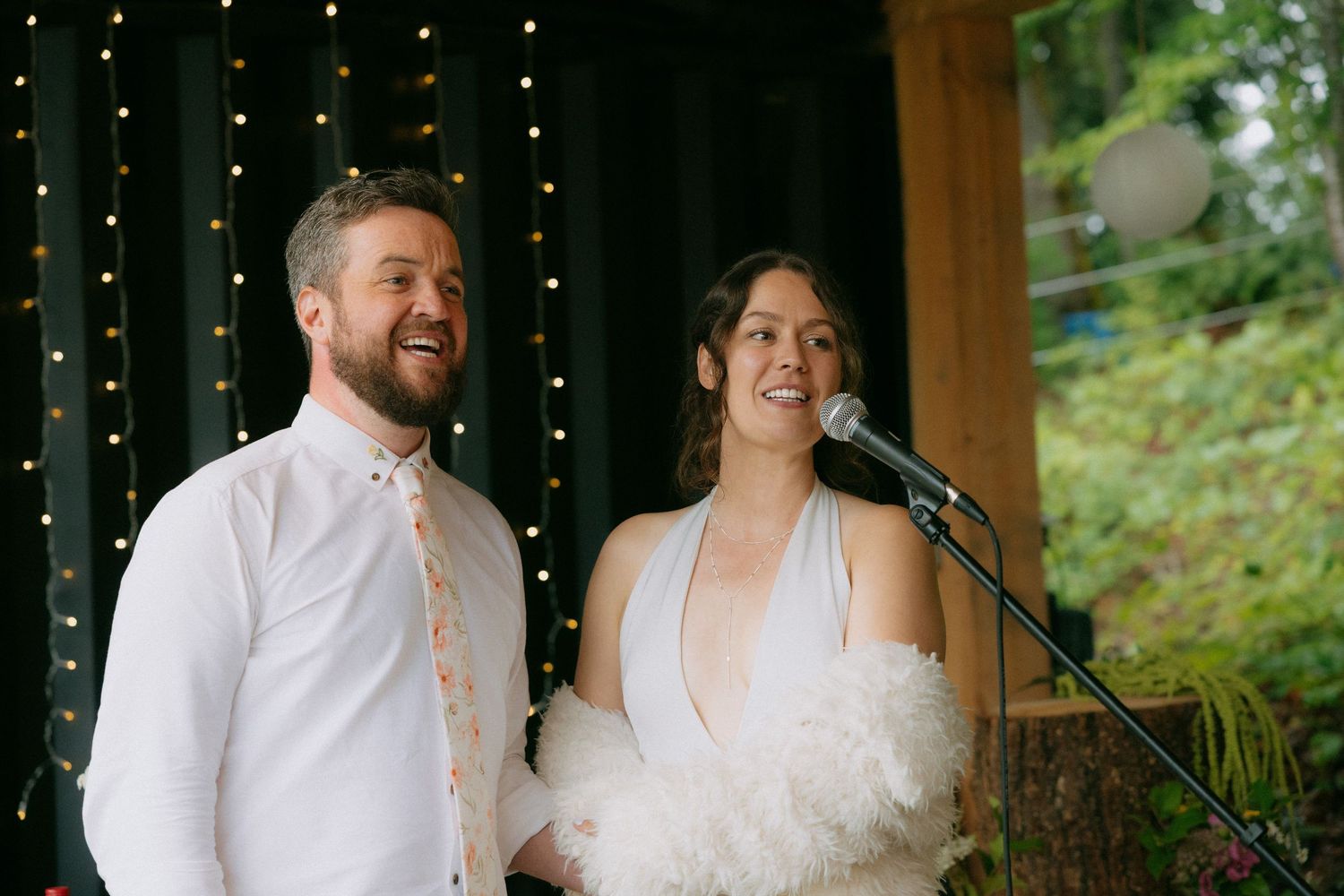 Happy couple smiling at microphone during wedding reception, bride in white halter gown with feathers, groom in white shirt with floral tie.