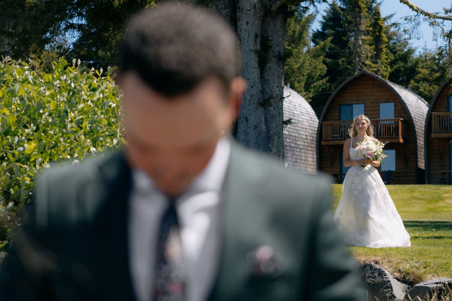 Bride in white gown holding bouquet stands near rustic cabin as groom in gray suit poses in foreground.