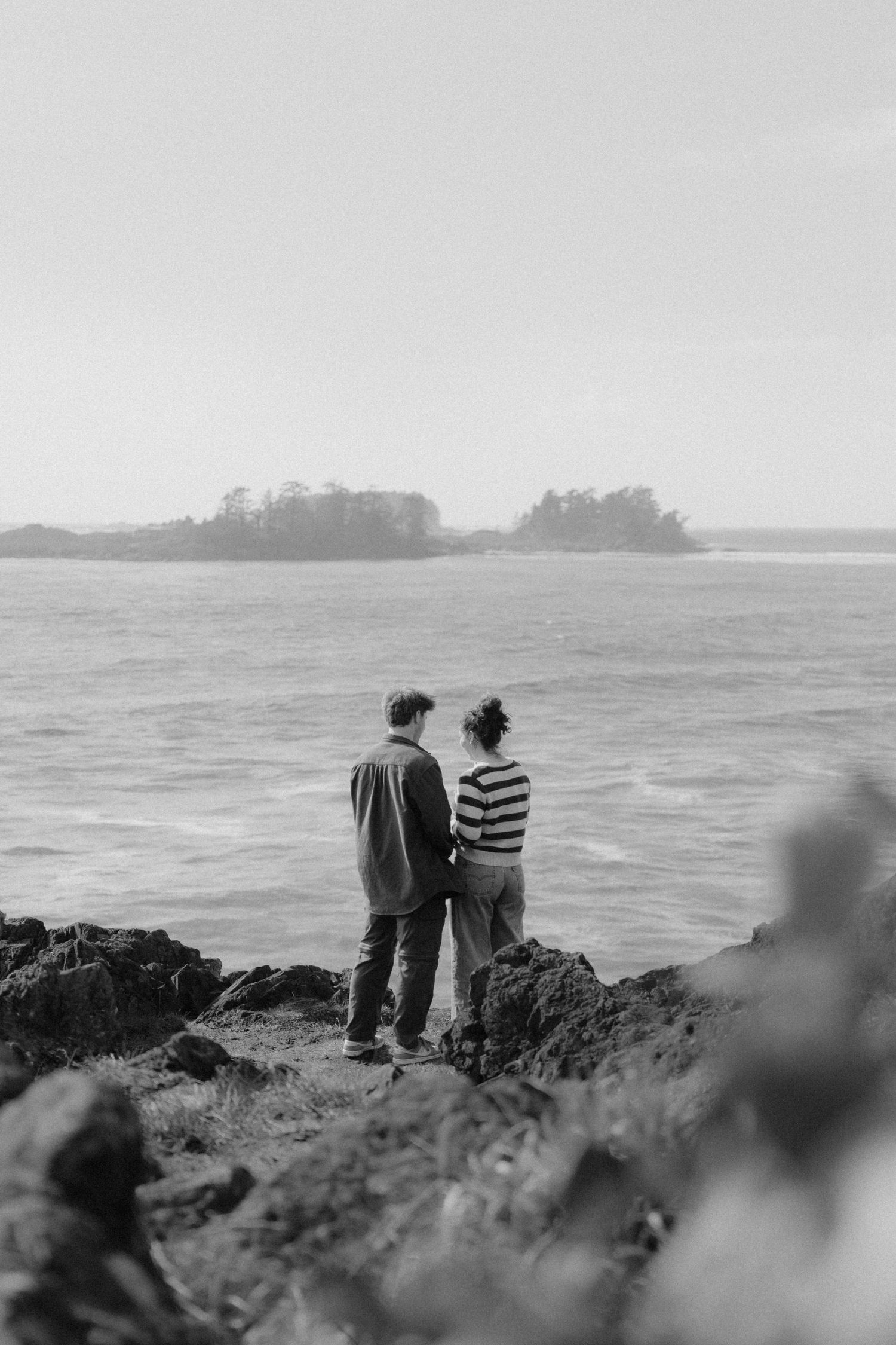Black and white photo of a couple standing on rocky cliffs overlooking the ocean with an island beyond.