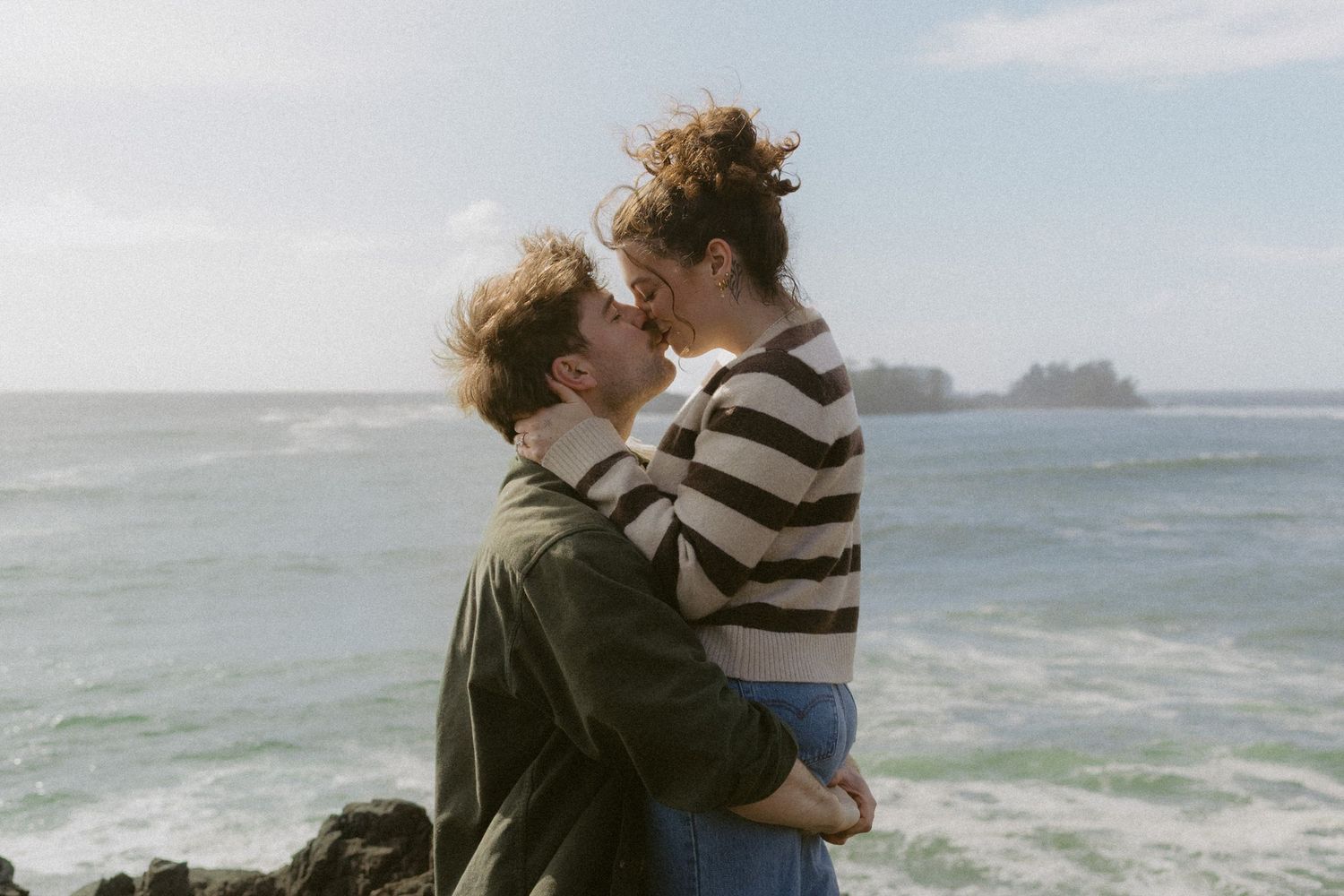 Romantic couple embracing and kissing on rocky seaside cliffs with ocean backdrop.