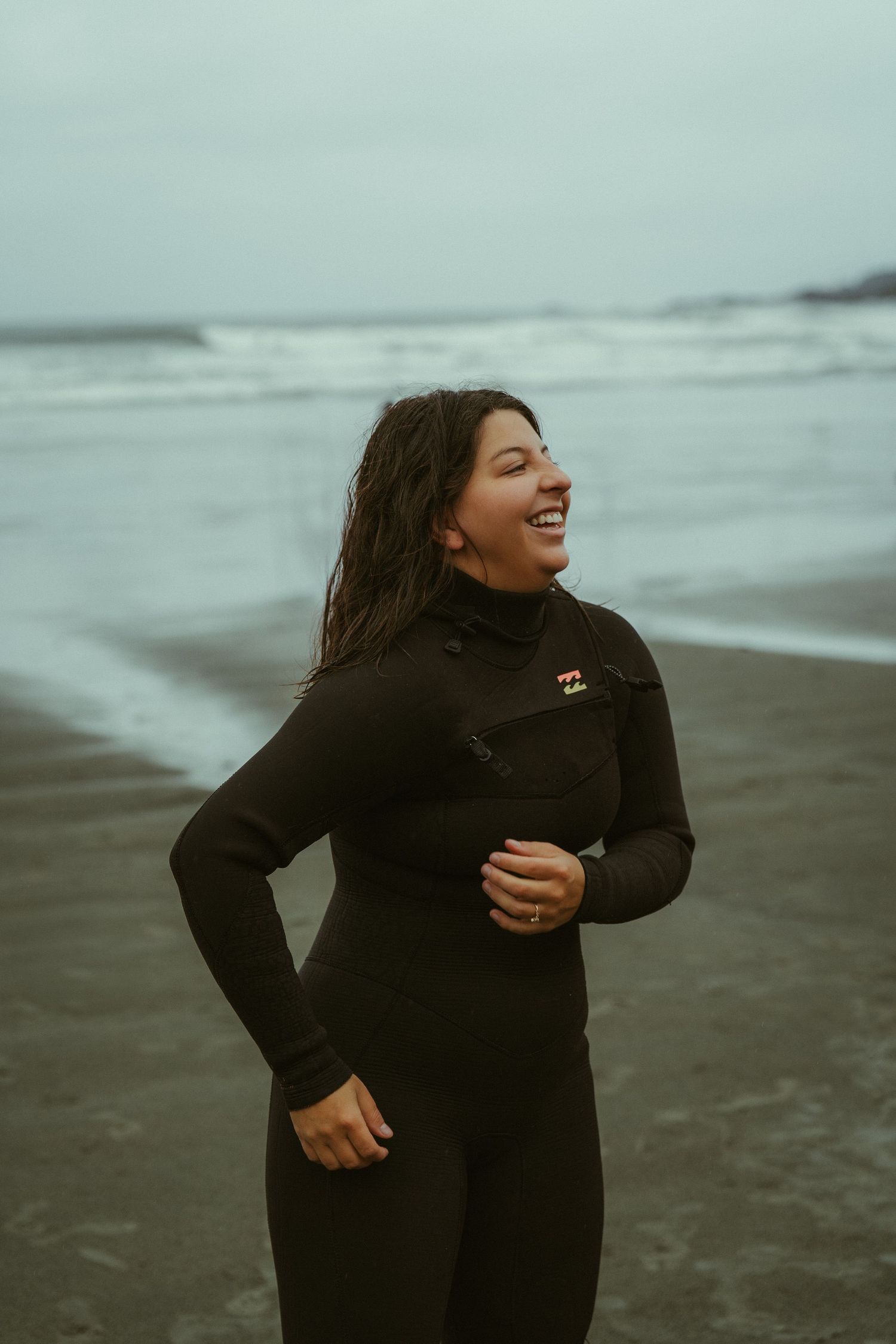 Port Alberni wedding photographer Mariana in black wetsuit smiling on cloudy beach on Cox Bay Tofino
