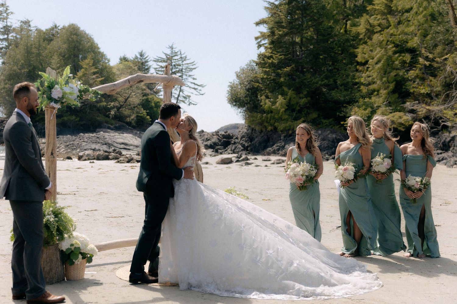 Couple shares first kiss at outdoor beach wedding ceremony with bridesmaids in sage green dresses and lush greenery.