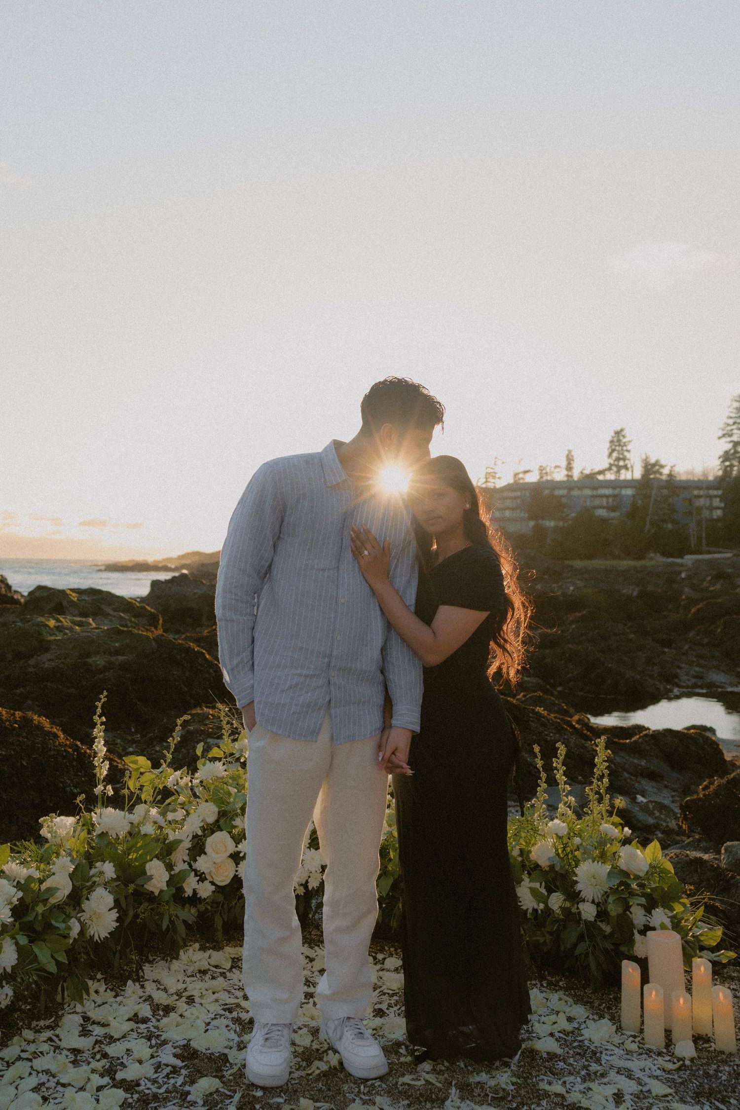Couple standing close together at sunset on rocky coast, surrounded by white flowers and candles.