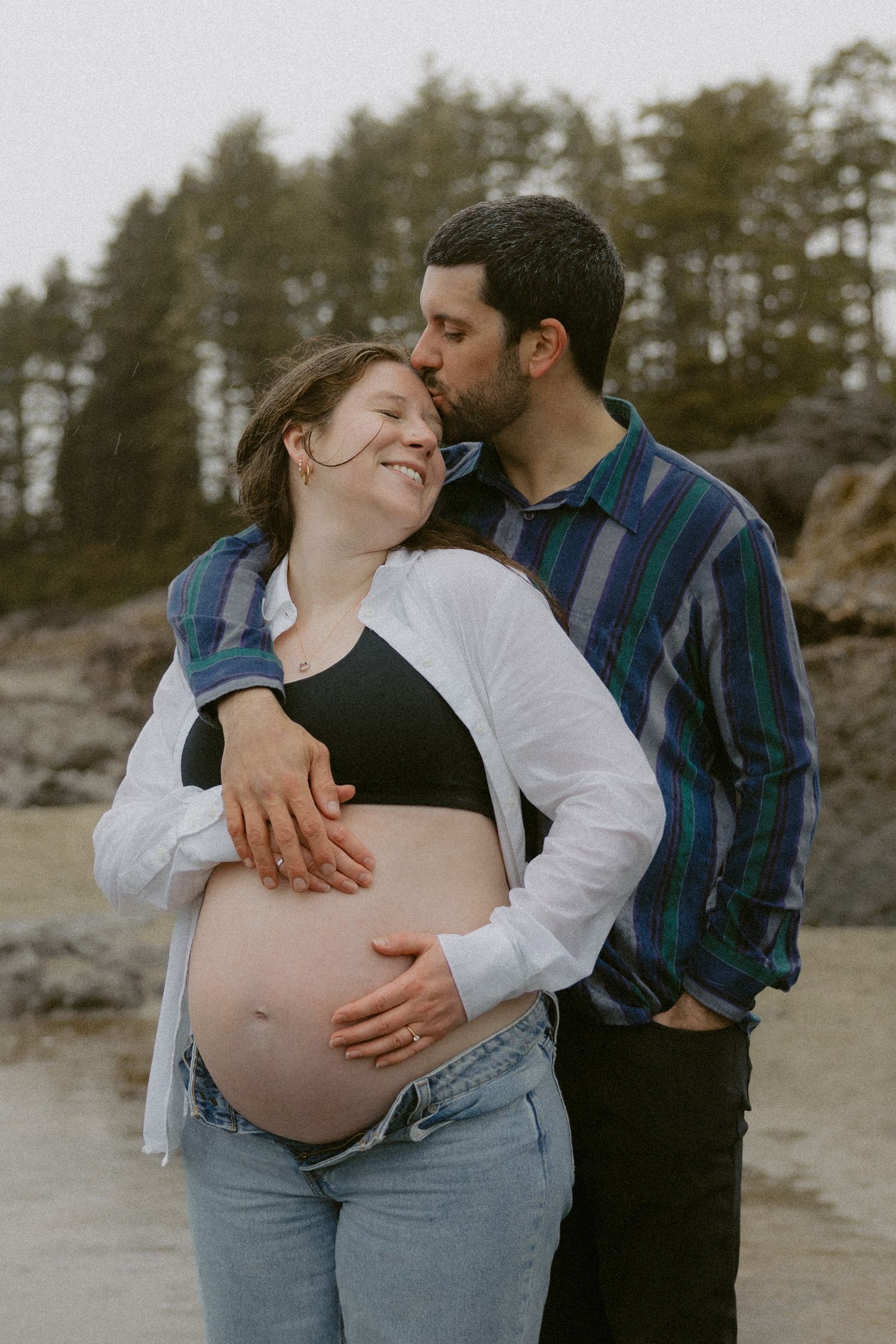 A couple shares an intimate moment on a rocky beach while showing off their pregnant belly.