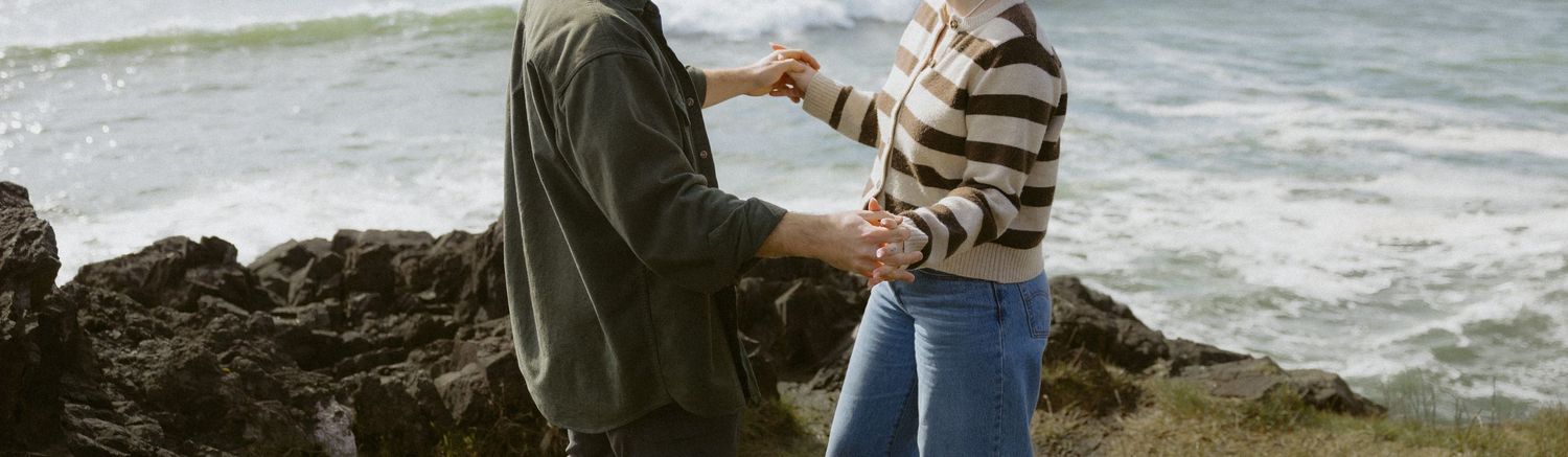 Couple holding hands near rocky coastline with ocean waves in background.