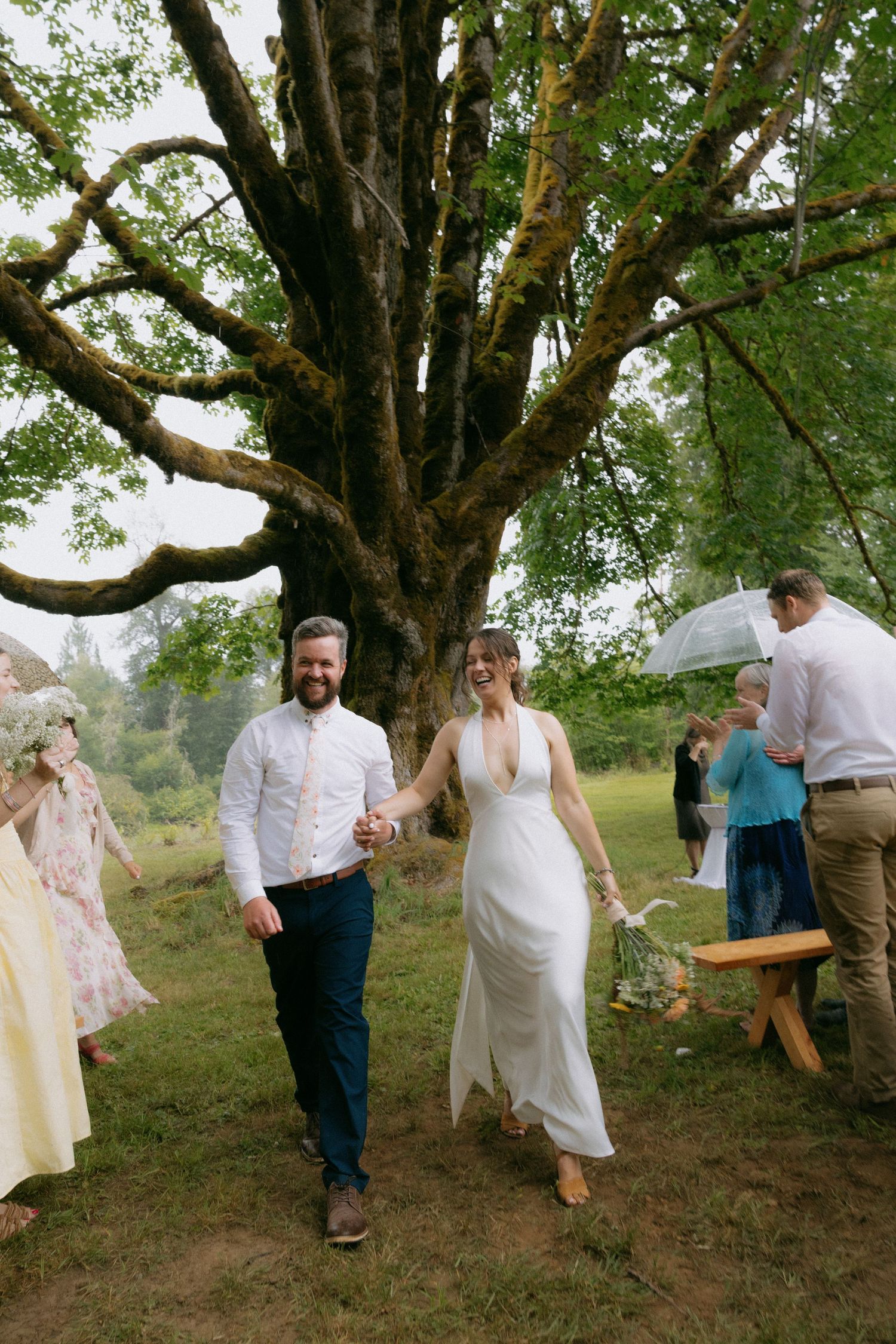 Happy bride and groom walking hand in hand after outdoor wedding ceremony beneath a large moss-covered tree.