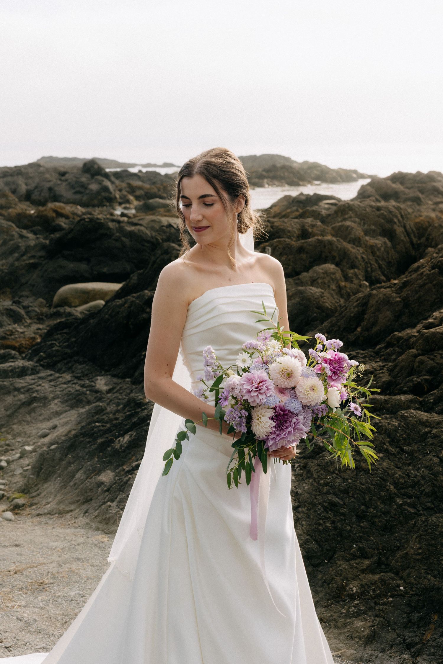 Bride in strapless white gown holding purple floral bouquet on rocky coastal beach.