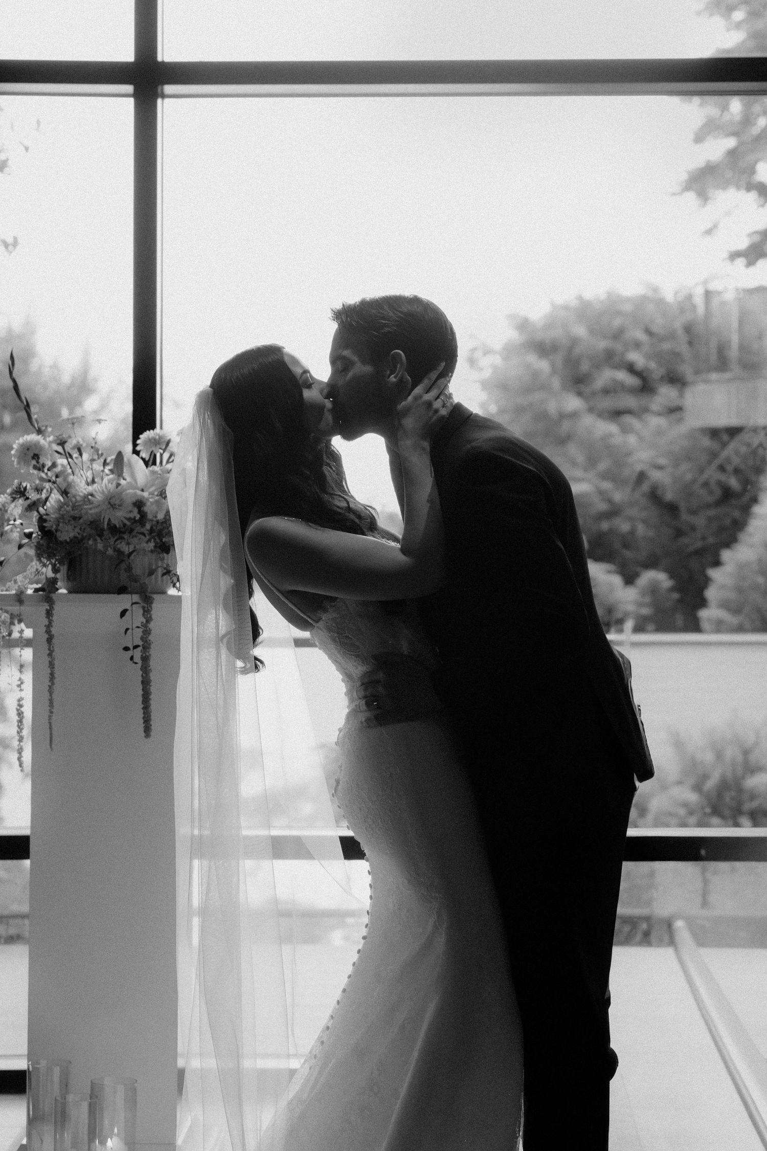 Black and white photo of a bride and groom sharing a kiss at the altar, with a flowing veil and floral arrangements.