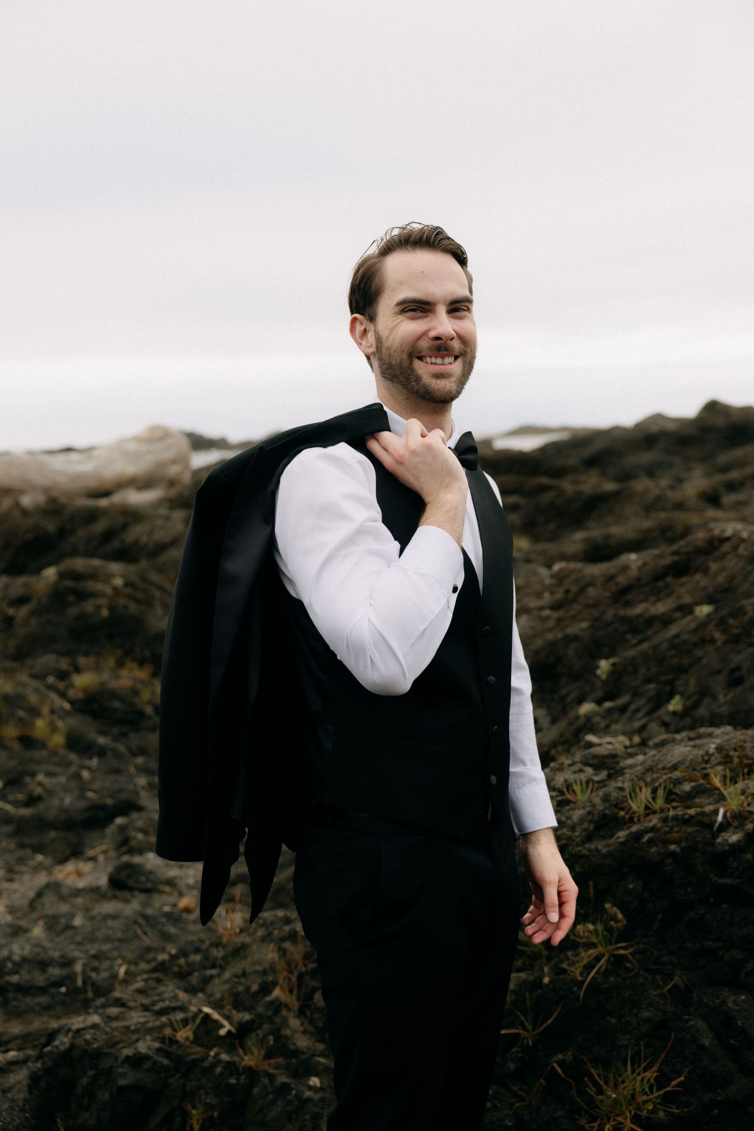 Smiling groom in black vest and bow tie holding jacket over shoulder on rocky outdoor landscape.