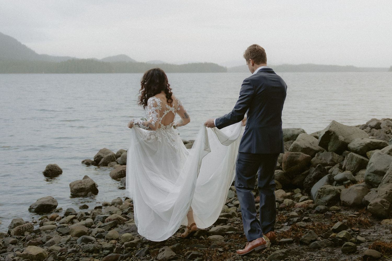 Two people explore a rocky beach shoreline with mountains and forest in the distance.