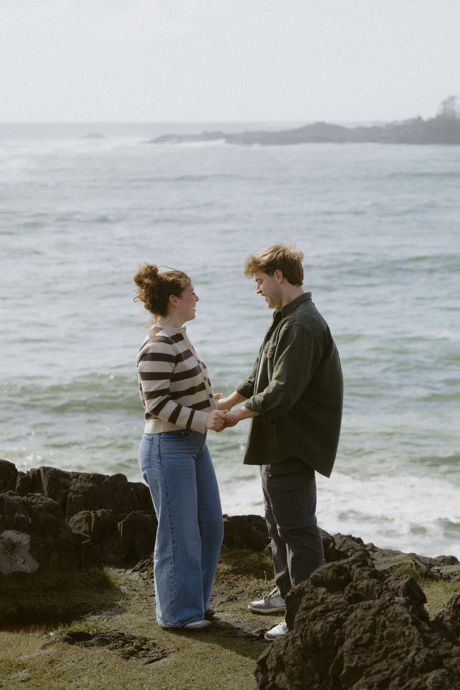 Couple holding hands facing each other on rocky beach with green ocean waves in background.