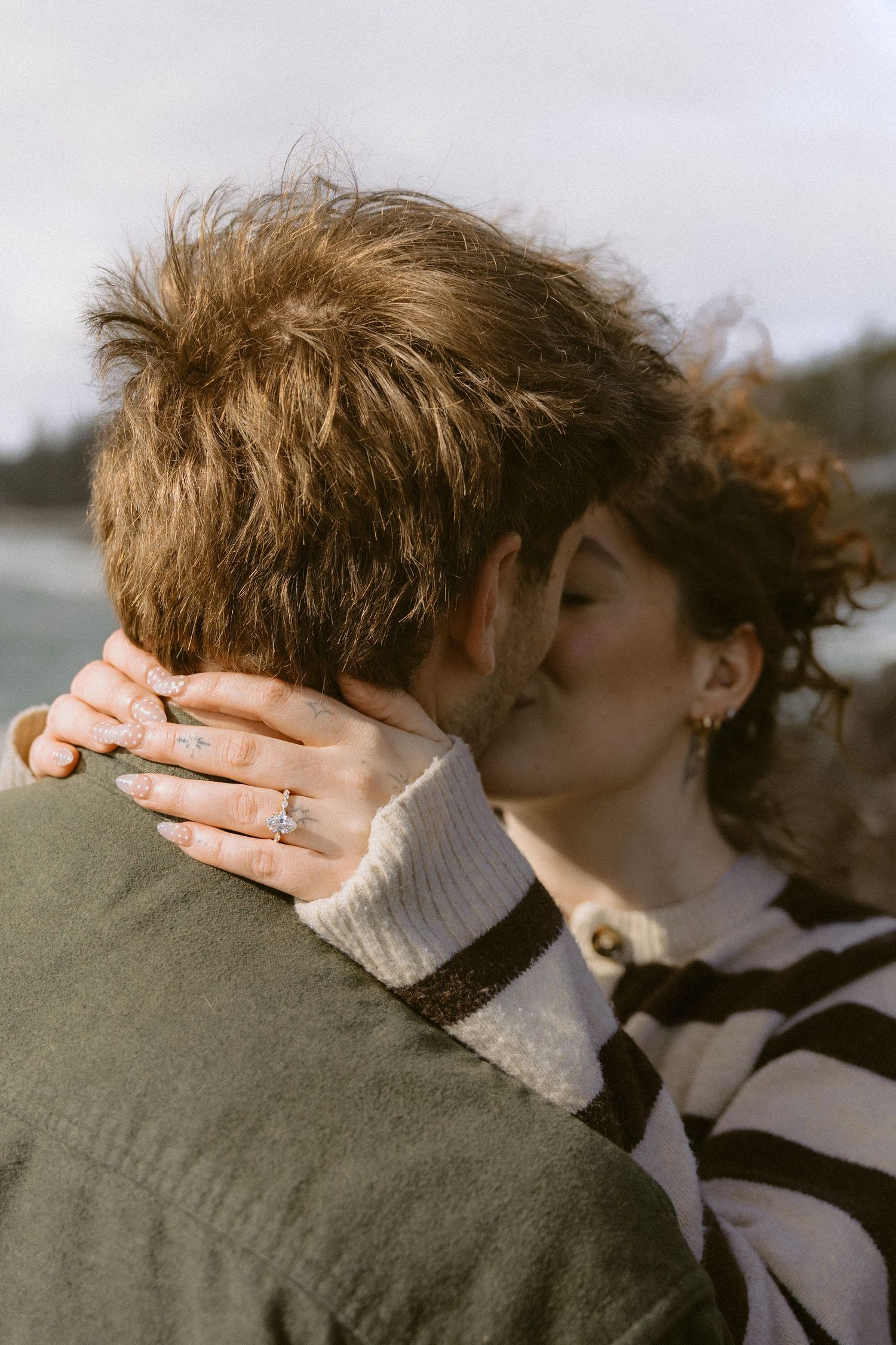 Couple sharing a romantic kiss outdoors, woman wearing an engagement ring with curly hair and striped sweater.