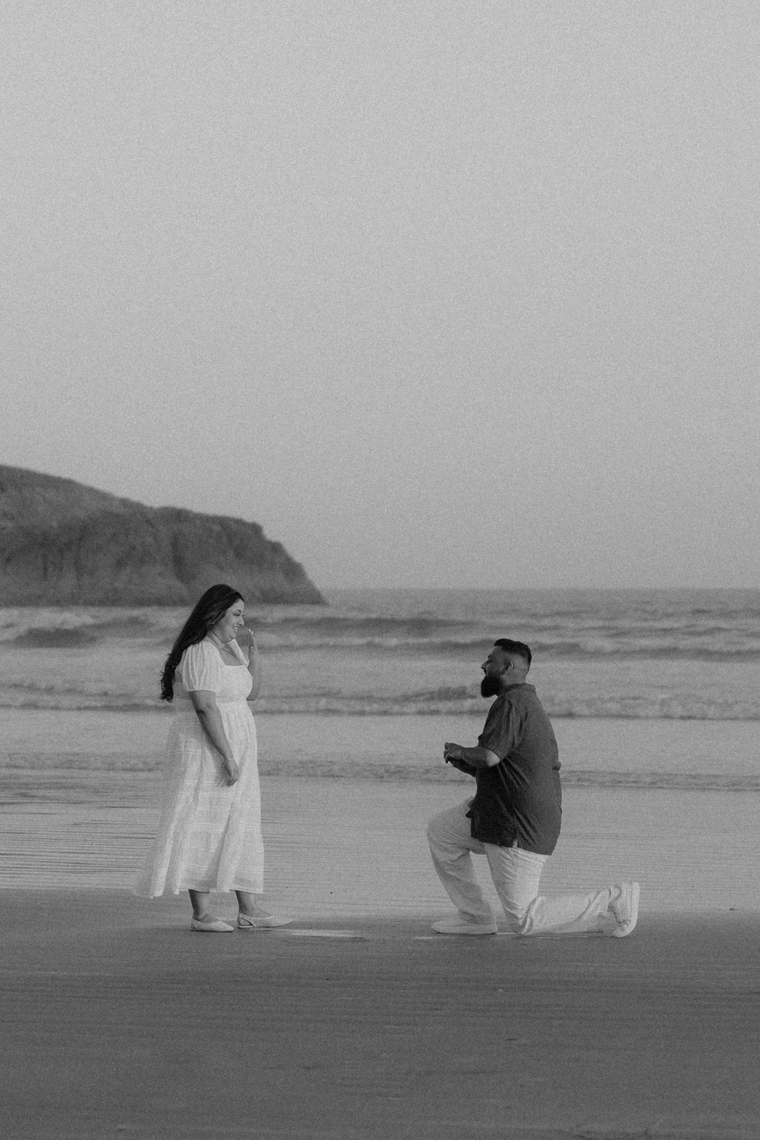 Black and white beach proposal photo of man on one knee proposing to woman in white dress at sunset by the ocean.