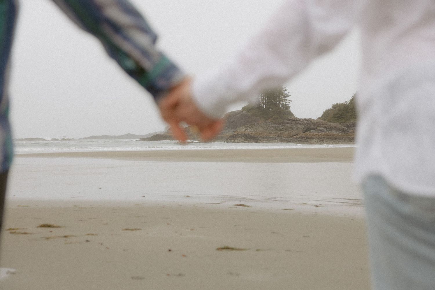 A blurred close-up shot of hands reaching toward each other on a misty beach with mountains in the background.