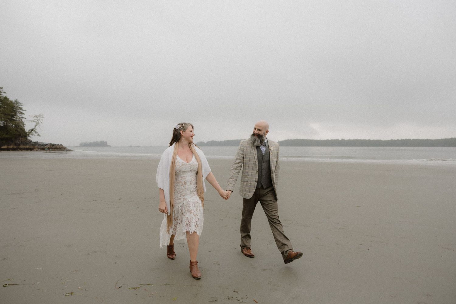 People walking along a foggy beach shoreline with evergreen trees visible in the background.