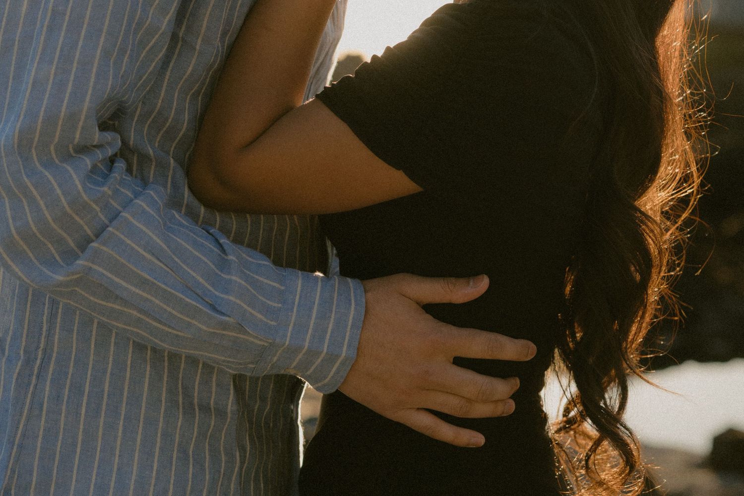 Couple embracing tenderly at golden hour, man in blue striped shirt holding woman in black dress close.