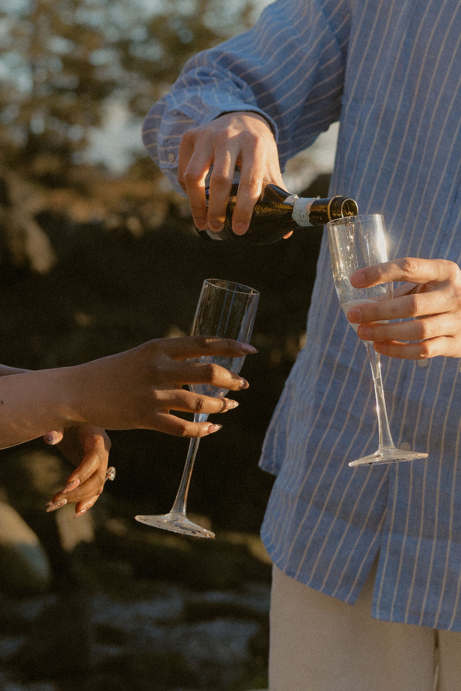 Person in blue striped shirt pouring champagne into crystal flutes outdoors at sunset.