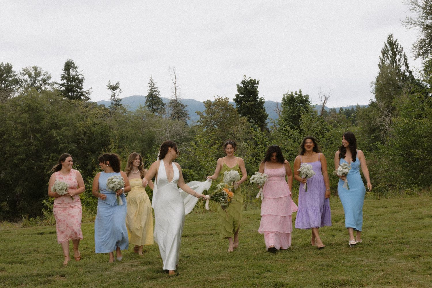 Bride in white gown walks with bridesmaids in colorful pastel dresses across a green meadow with mountain views.