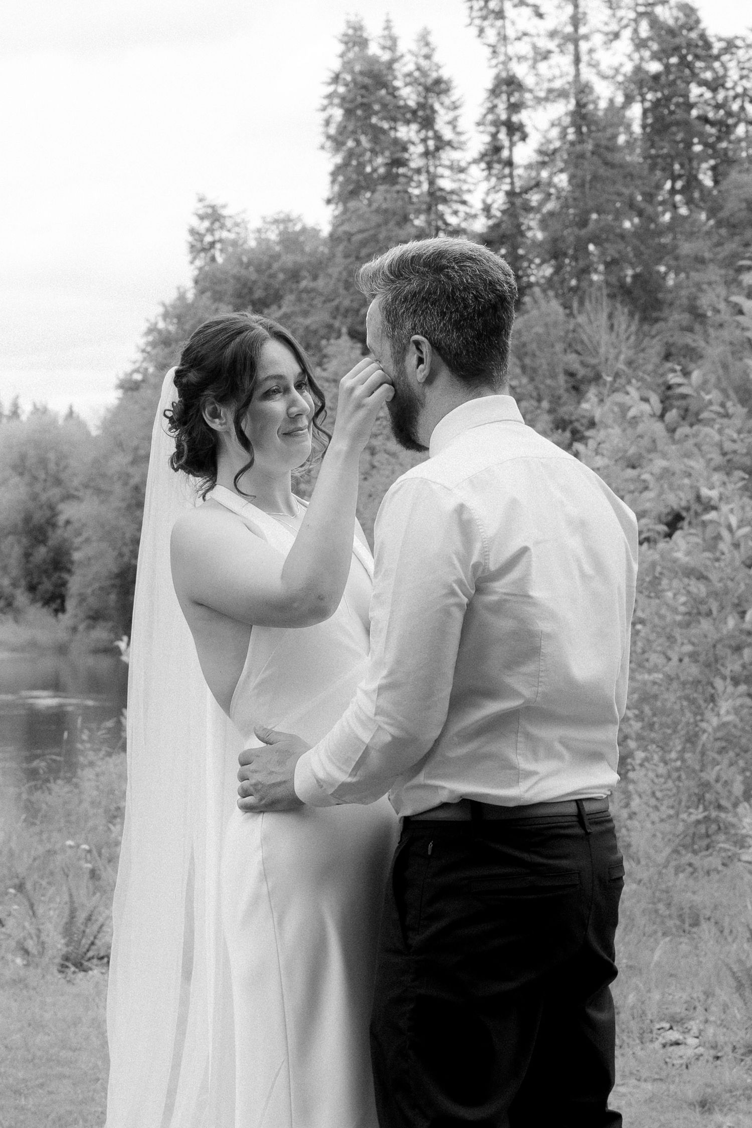 Black and white photo of a smiling bride in a veil touching the groom's face outdoors near trees and a pond.