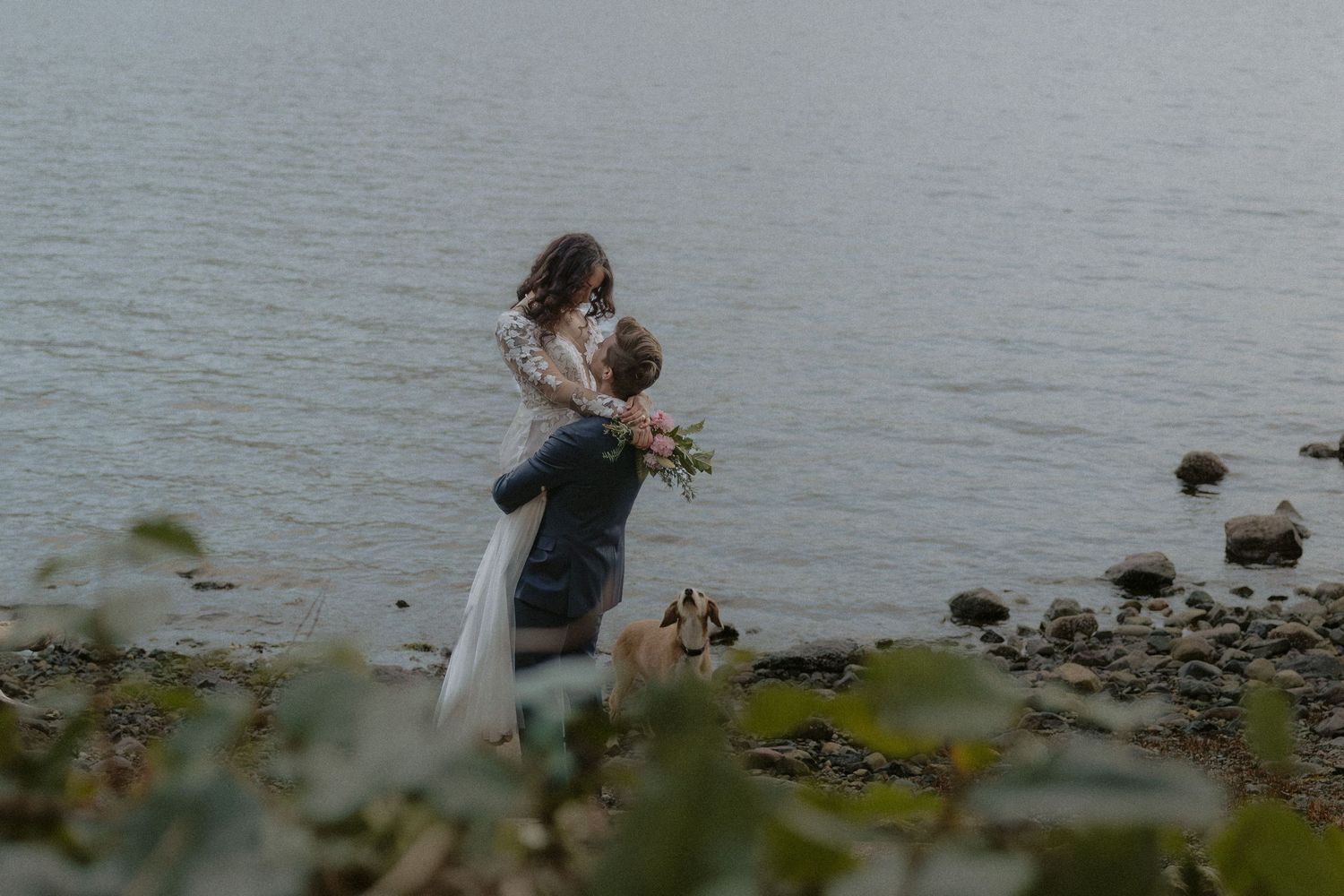 Couple walking along a misty waterfront in romantic wedding attire.