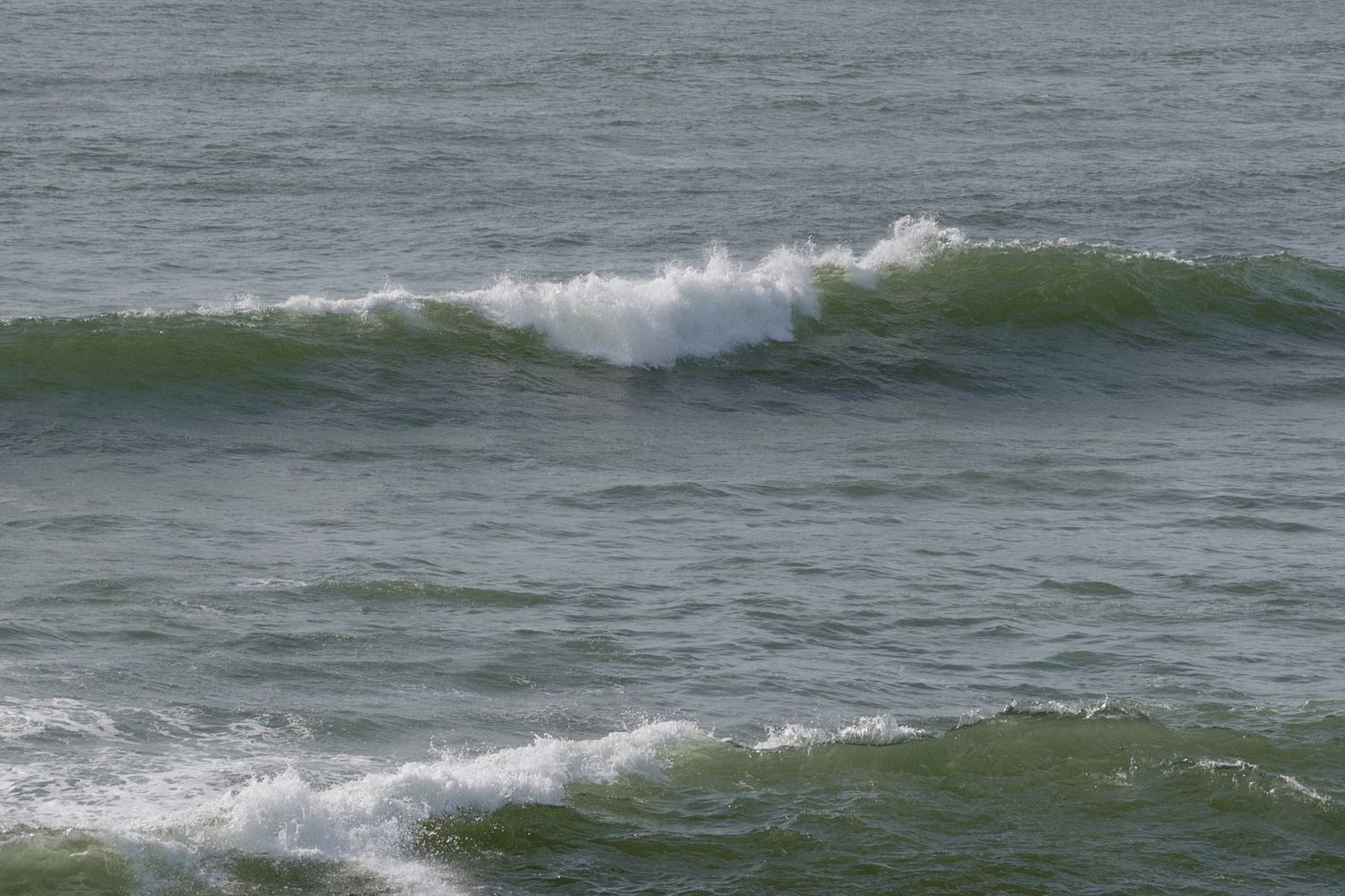 Ocean waves with white foam cresting on green-blue water near a sandy shore on a calm, overcast day.