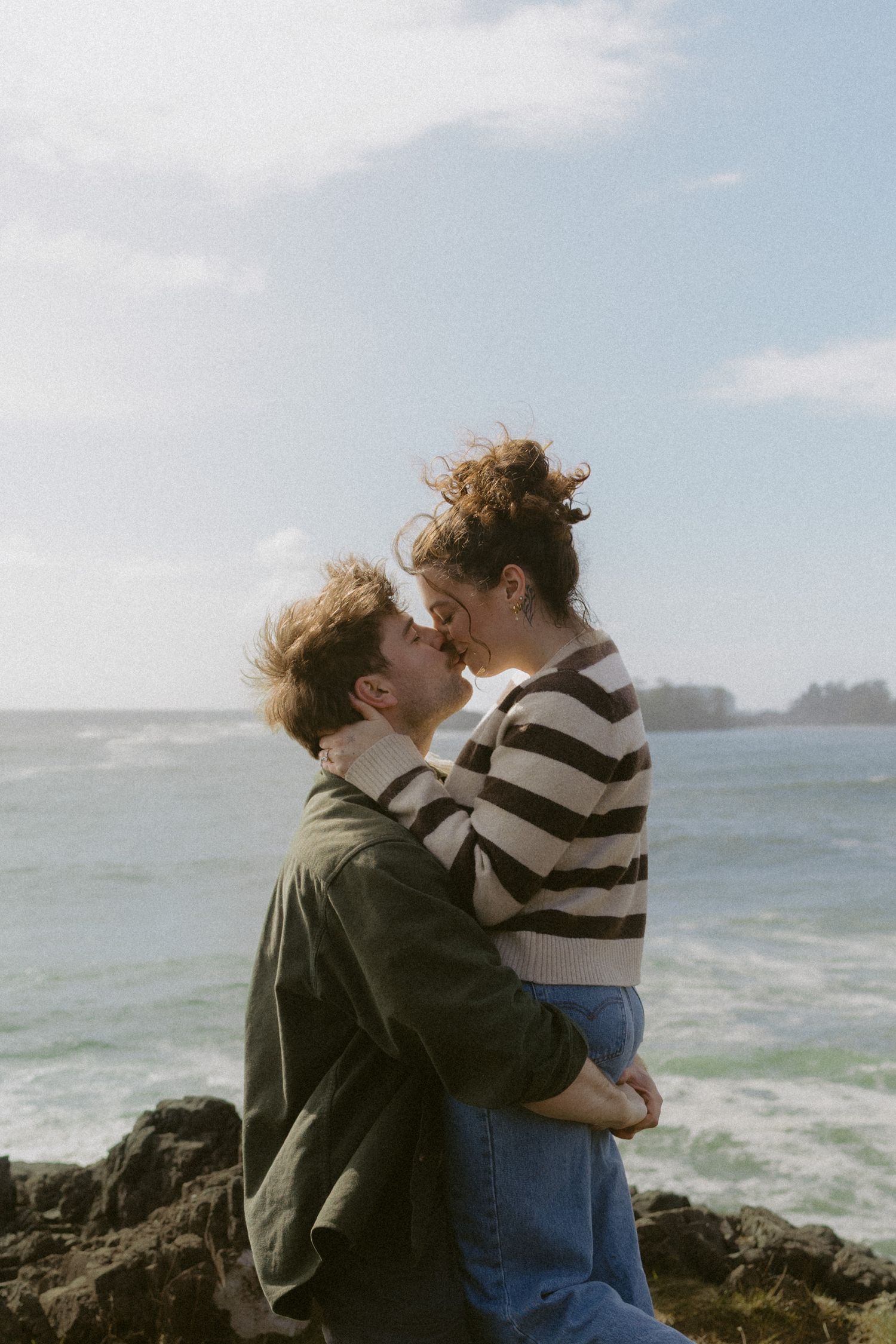 Couple sharing a romantic kiss on rocky coastal cliffs with ocean waves behind them.