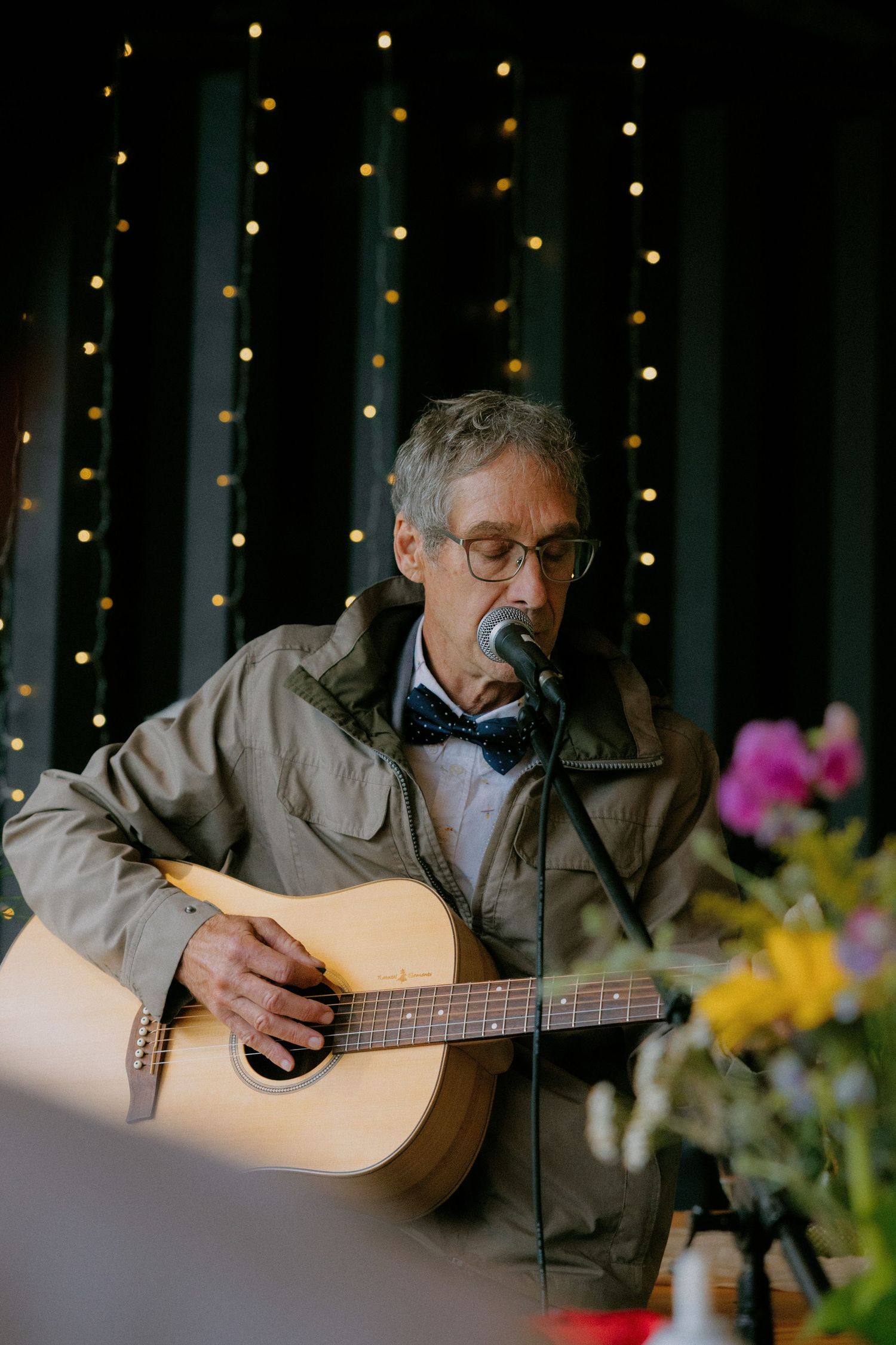 Older musician with glasses and bow tie plays acoustic guitar at a wedding reception.
