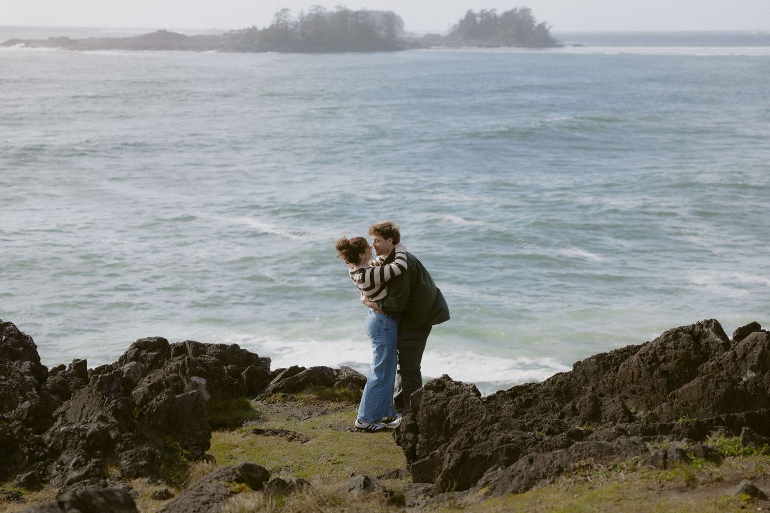 Couple embracing on rocky coastal cliffs with ocean waves and misty green island in the background.