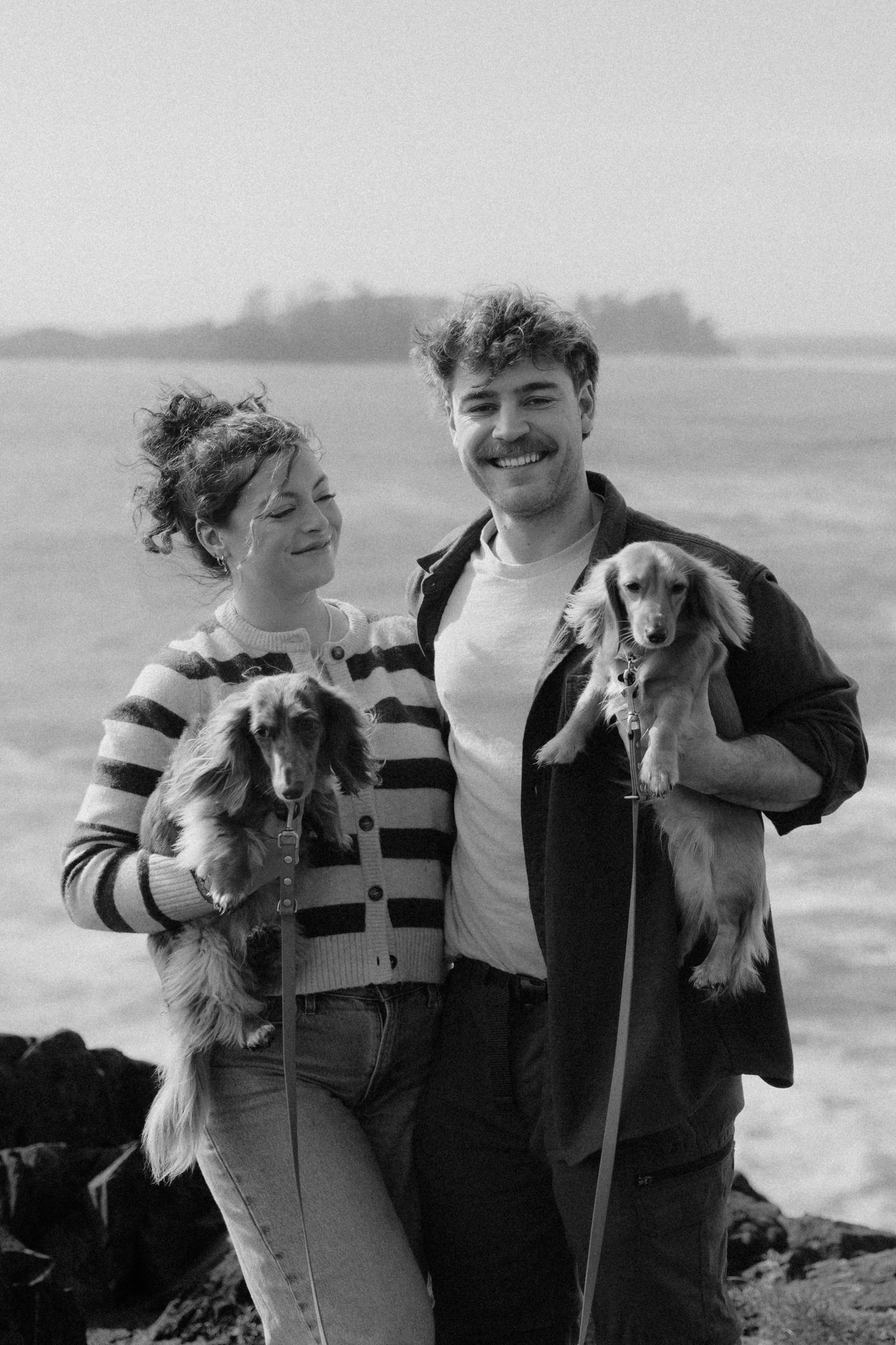 Smiling couple holding two golden dachshund dogs on rocky beach in black and white photo.