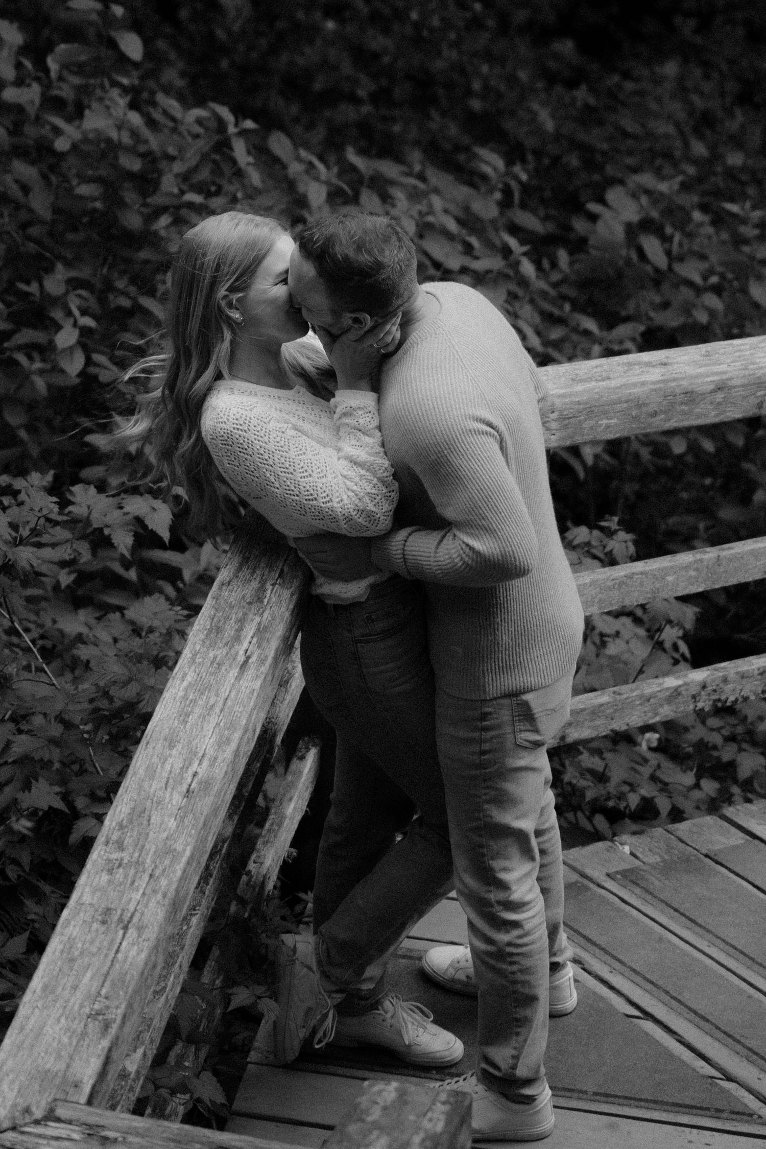 Black and white photo of a couple kissing on a wooden bridge surrounded by lush green foliage.