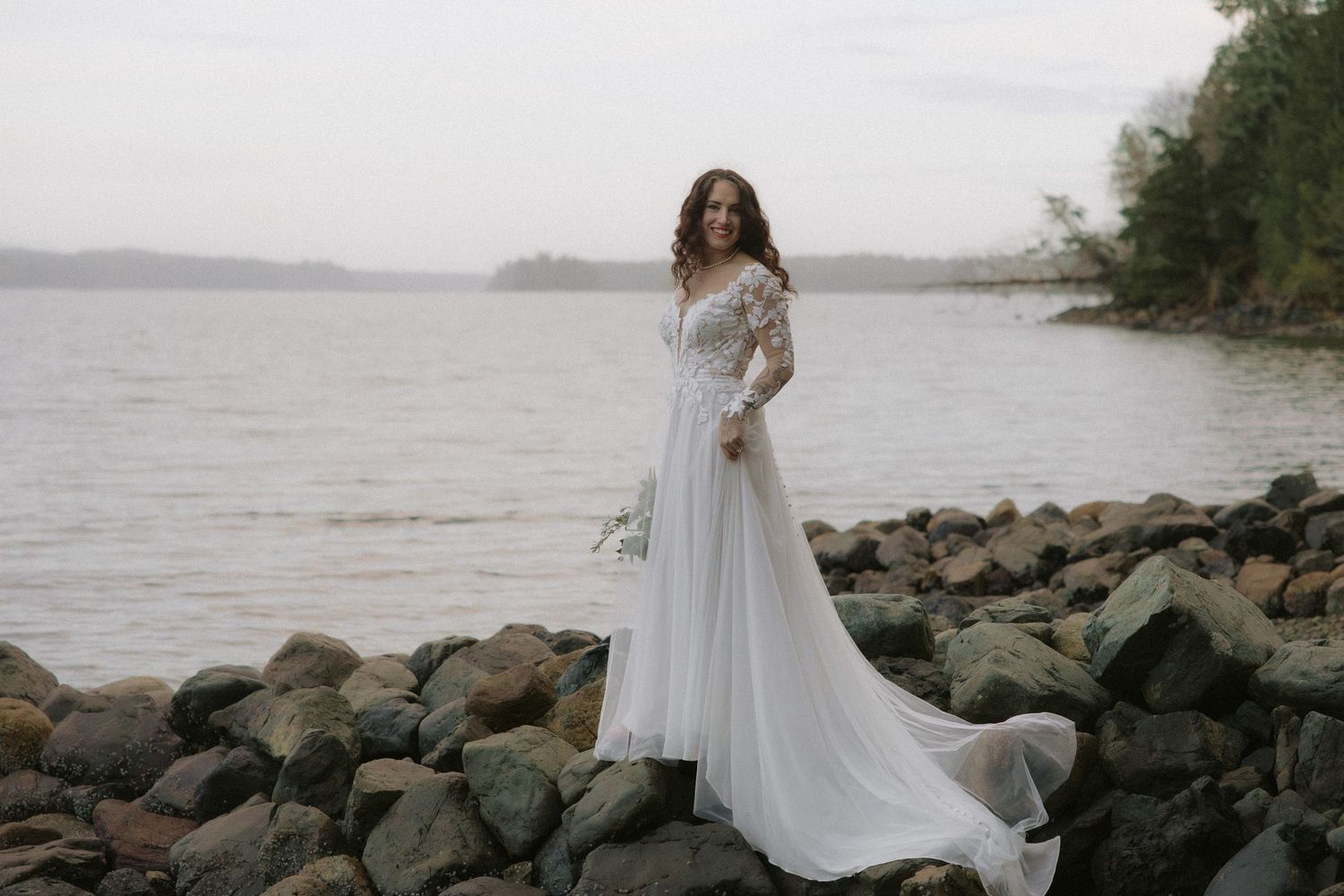 A romantic wedding photoshoot on a rocky beach with mountains and water in the background.