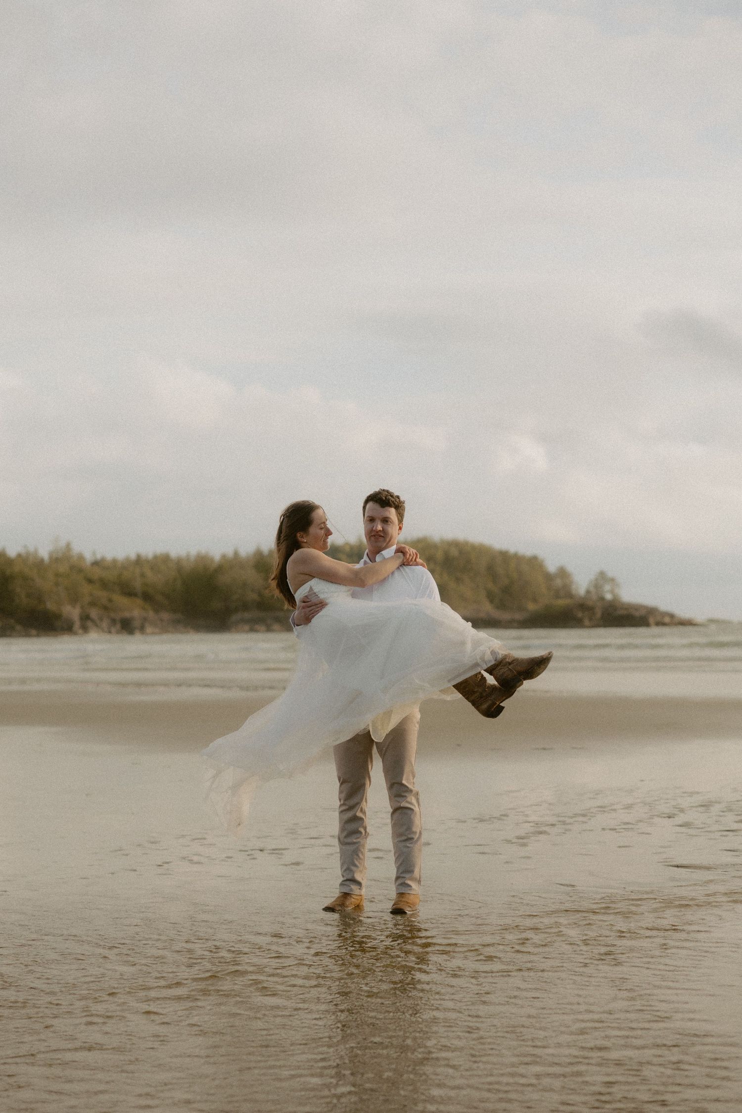Groom carries bride across shallow beach water during sunset